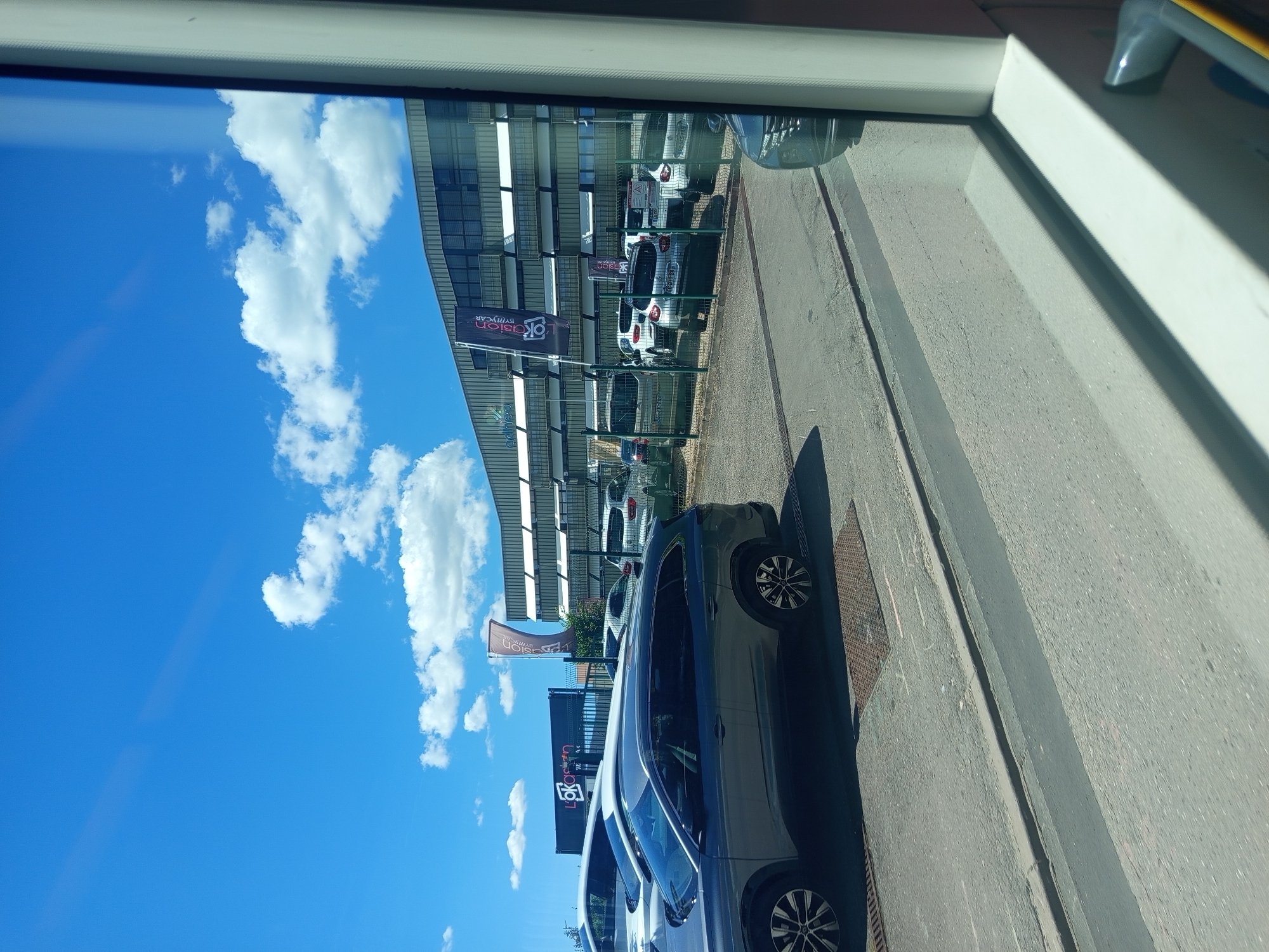 The image depicts a car dealership lot in Laxou, France, on a sunny day with a bright blue sky and scattered white clouds. In the foreground, a dark grey SUV is parked, facing left. Behind it, several other cars are lined up, including white and silver models. A modern, multi-story building with large windows and a metallic facade serves as the backdrop. Banners with the "l'okasion" logo are visible on flags and the building. The scene is viewed from inside a vehicle, with a window frame partially obscuring the view on the right side. The ground is paved, with a metal grate visible in the foreground.