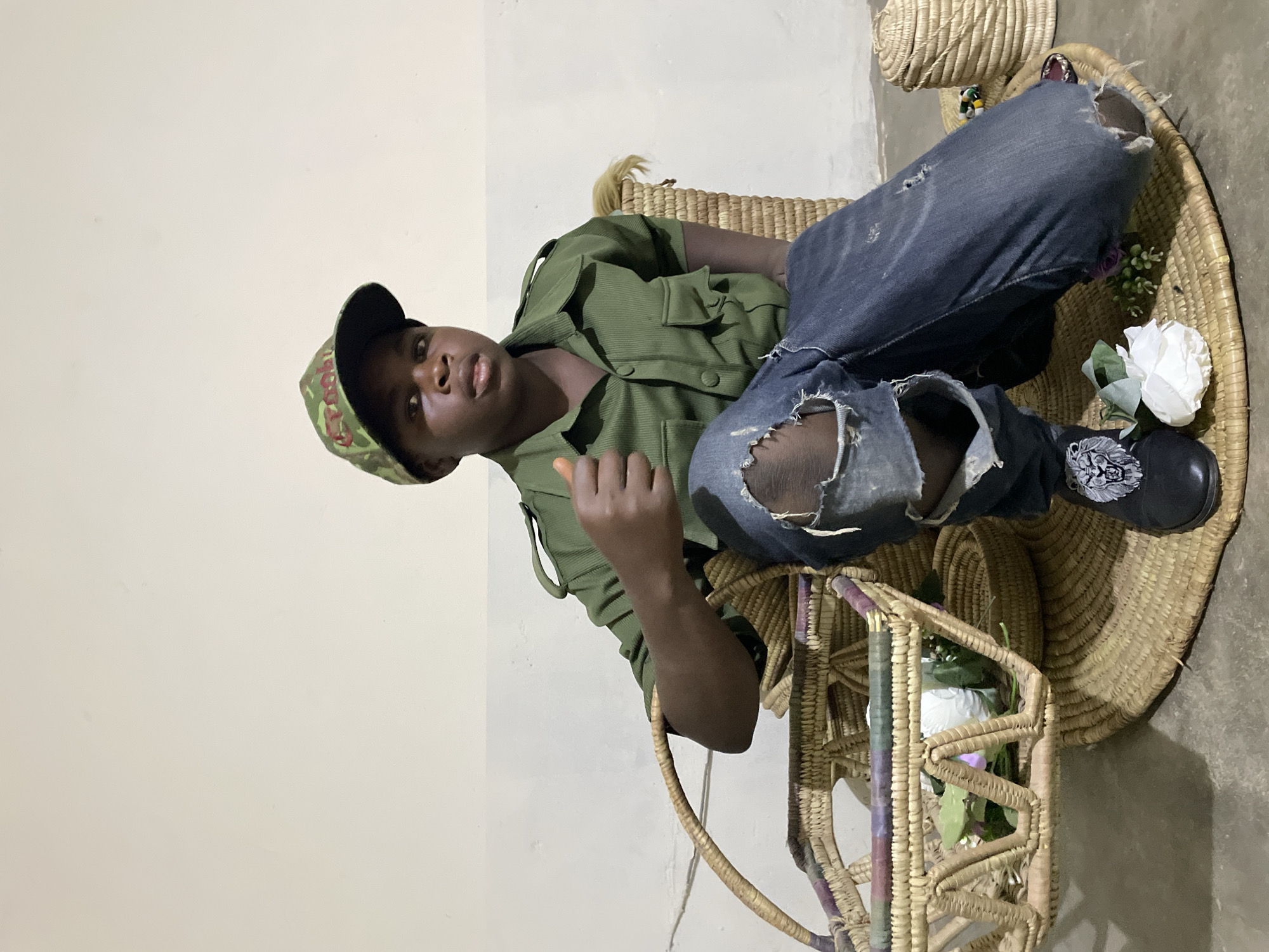 A young man is sitting in a woven chair. He is wearing a green button-down shirt, blue jeans with a large tear in the right knee, a black baseball cap with a camouflage pattern, and black shoes. The chair is woven, and there are white flowers in a basket near the bottom of the chair. There is a woven basket behind the chair and another to the right. The background is a plain white wall and a gray floor. The location is Jos, Nigeria.