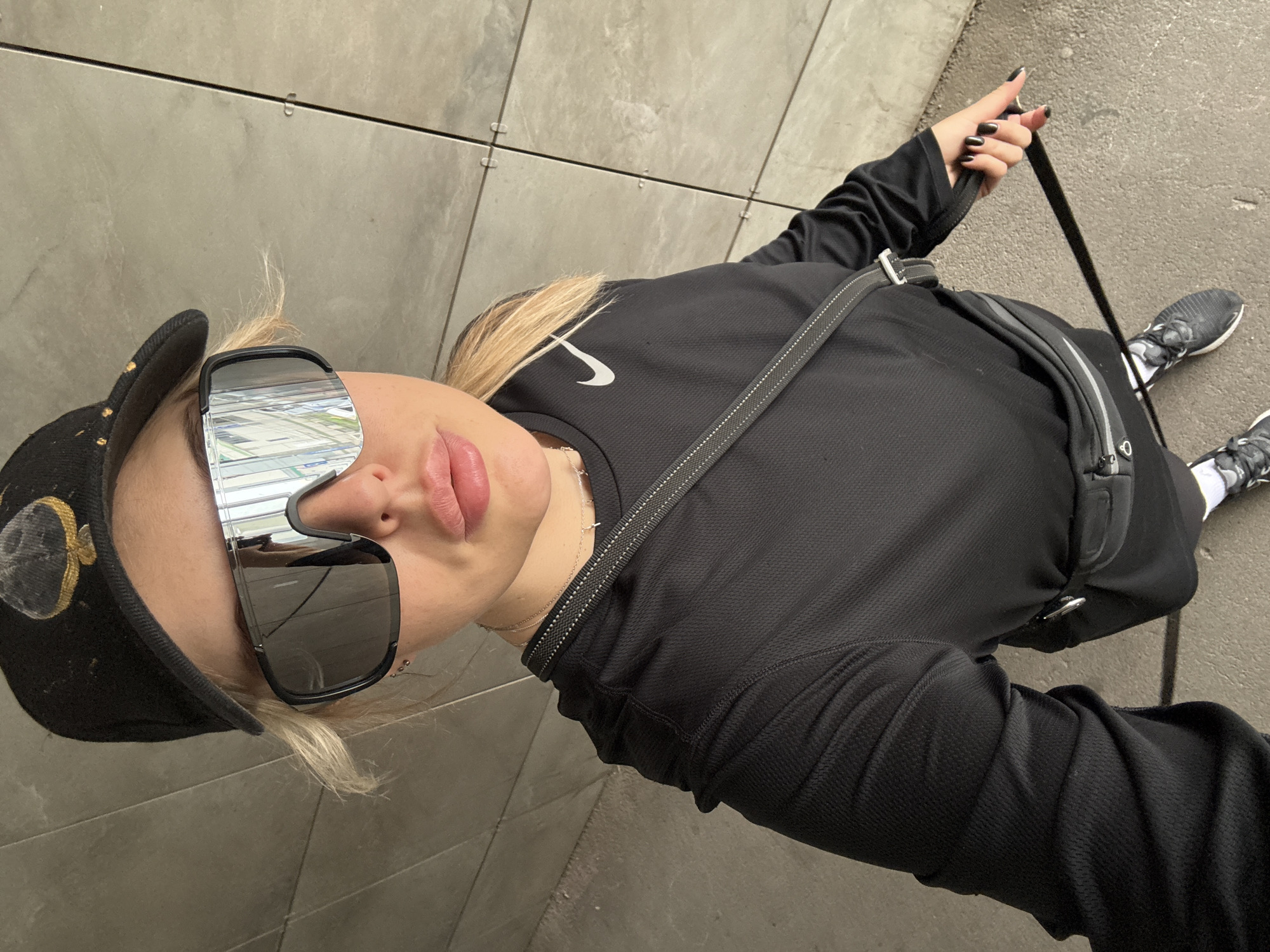 A woman wearing a black cap with a gold design, large sunglasses reflecting buildings, and a black Nike long-sleeved shirt and pants is on a city street. She has blonde hair and wears a silver necklace. She holds a black leash in her right hand, and wears black running shoes. The environment consists of tiled sidewalks and asphalt. Based on the reflected buildings, the location is likely Krasnoyarsk, Russia.