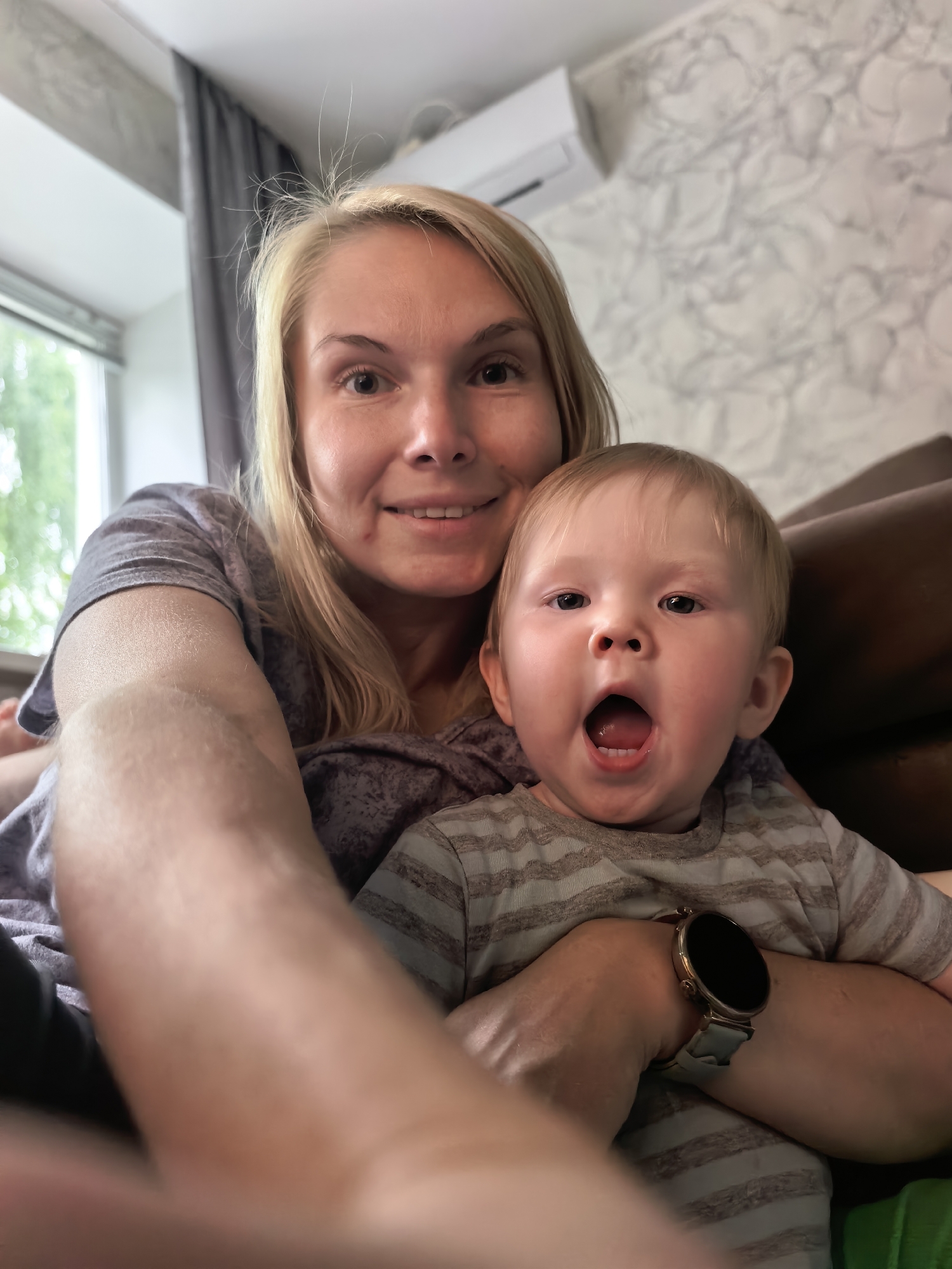 A woman with blonde hair smiles while holding a baby on her lap. The baby has blond hair and is looking at the camera with an open mouth. The woman is wearing a purple shirt, and the baby is wearing a striped shirt. A smartwatch is visible on the woman's wrist. The background appears to be an indoor setting with a window and a wall with a textured pattern.