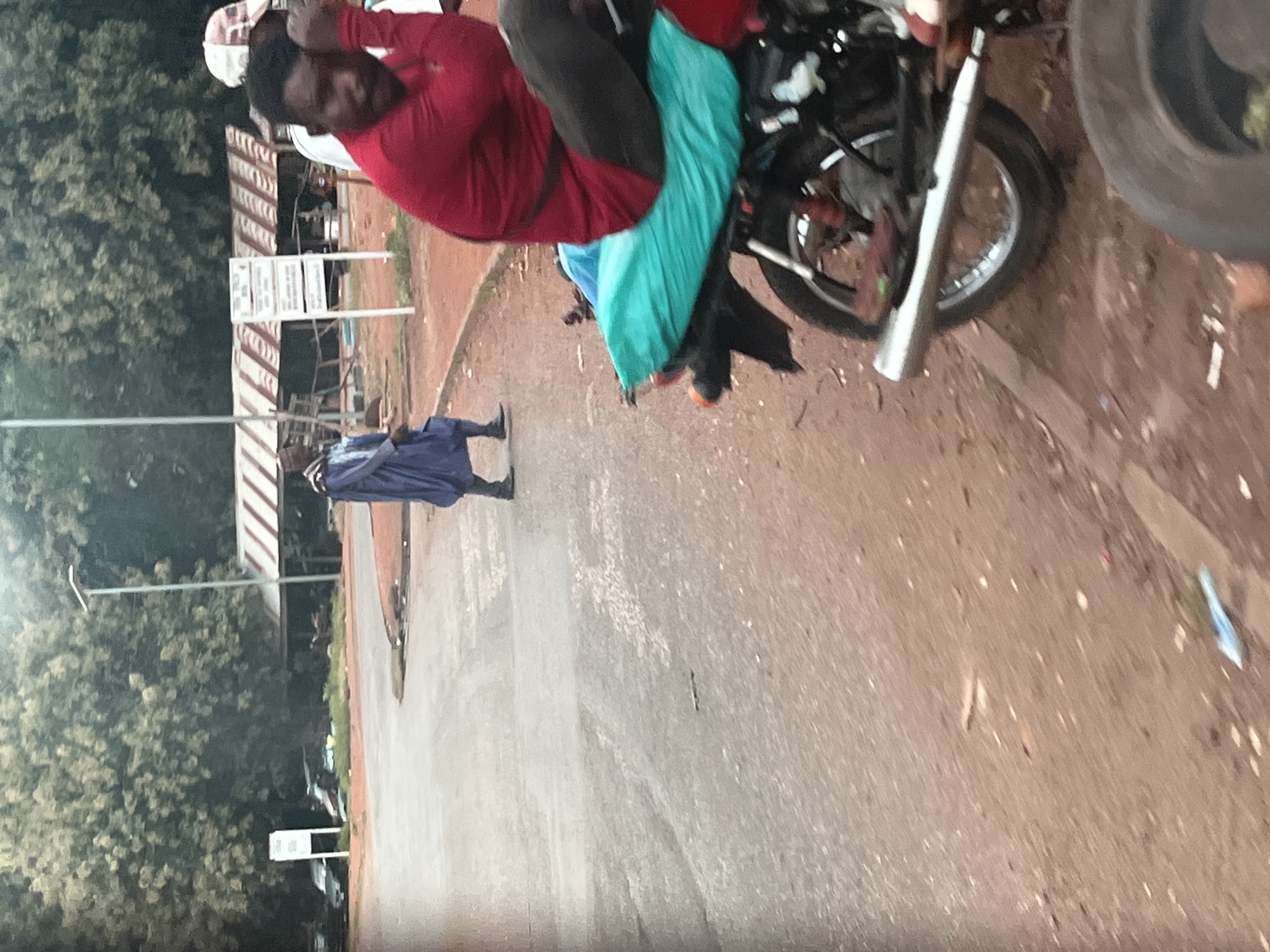 In the city of Gidan Waya, Nigeria, a person dressed in a blue robe and a traditional cap walks on a paved road. A man in a red shirt and dark pants sits on a motorcycle that is parked on the side of the road, with a teal-colored cover on the seat. The motorcycle has a silver exhaust pipe and a visible rear wheel. In the background, trees and a structure with a striped roof are visible, along with a signpost. The environment appears to be outdoors, with a dirt shoulder bordering the paved road.