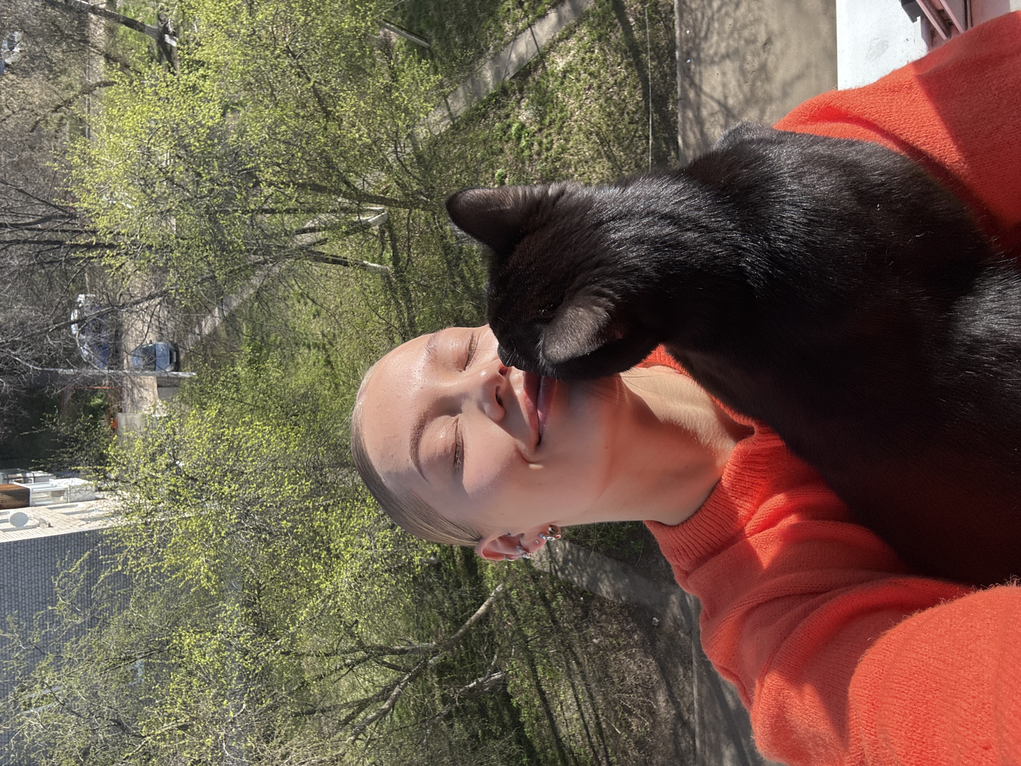 A woman wearing an orange sweater is standing outside with a black cat, which appears to be licking her face. The location is Chaikovsky, Russia. Trees and buildings are visible in the background, suggesting an urban environment. The photo is taken during daylight, under sunny conditions.