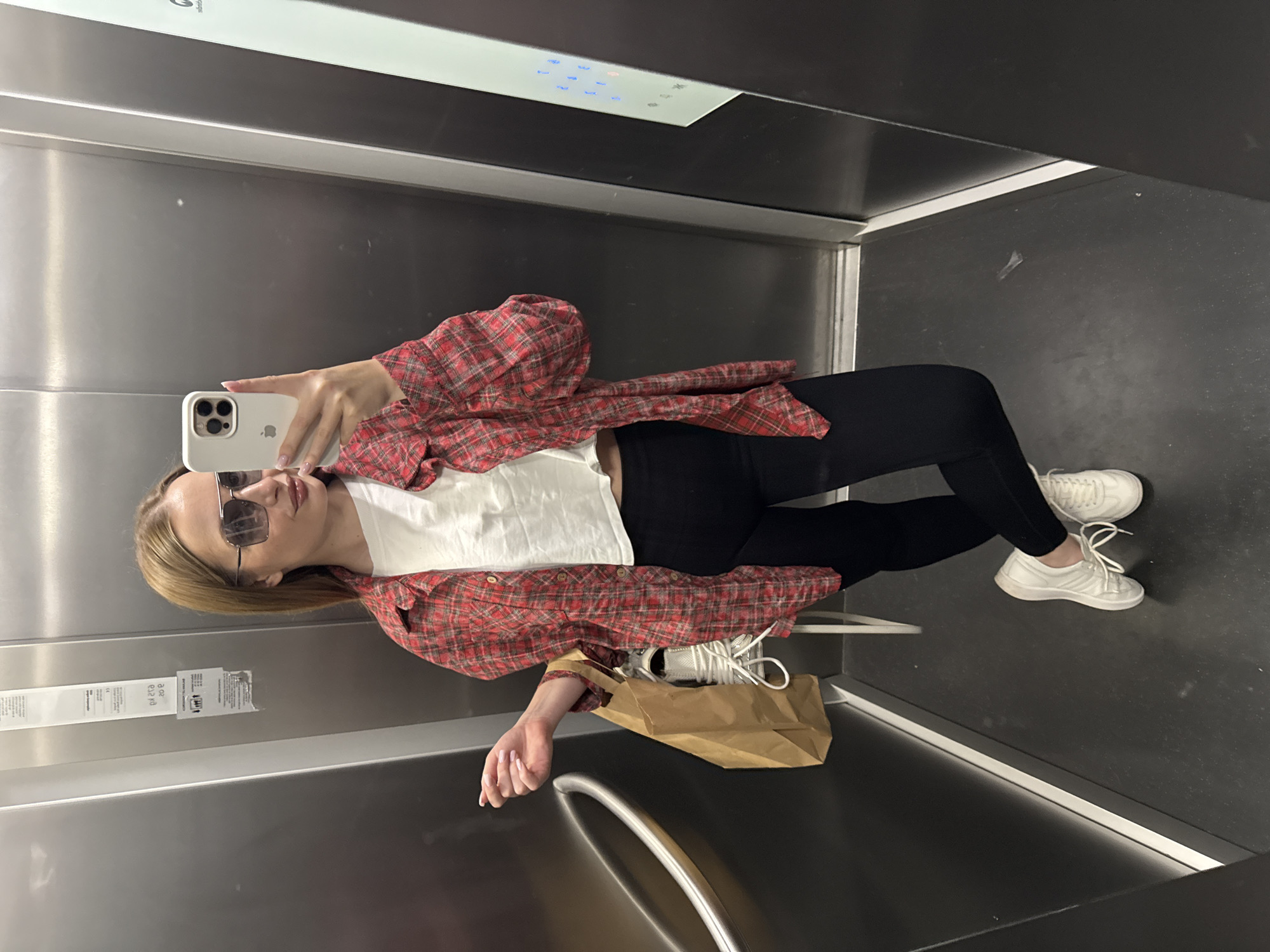 The image shows a woman taking a selfie in an elevator. The elevator has stainless steel walls and a black floor. She is wearing sunglasses, a white t-shirt, a red plaid shirt, black leggings, and white sneakers. She is holding a white smartphone in her right hand. A brown paper bag sits on the floor beside her feet. Based on the sticker affixed to the inside of the elevator, the elevator is located in Budva, Montenegro.