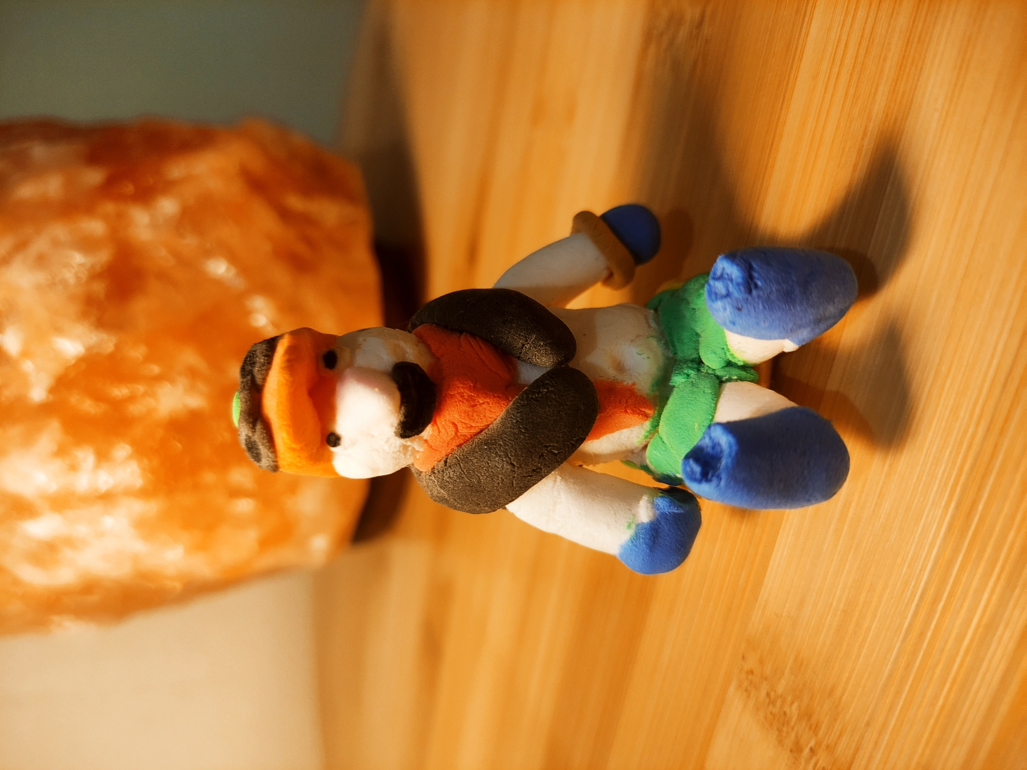 A clay figure lies on its side on a wooden surface, with an orange object blurred in the background. The clay figure has an orange hat, white face, black mustache, orange vest, black belt, white arms, green pants, and blue boots and gloves. The wooden surface has vertical grains. The blurry orange object is to the left of the clay figure.