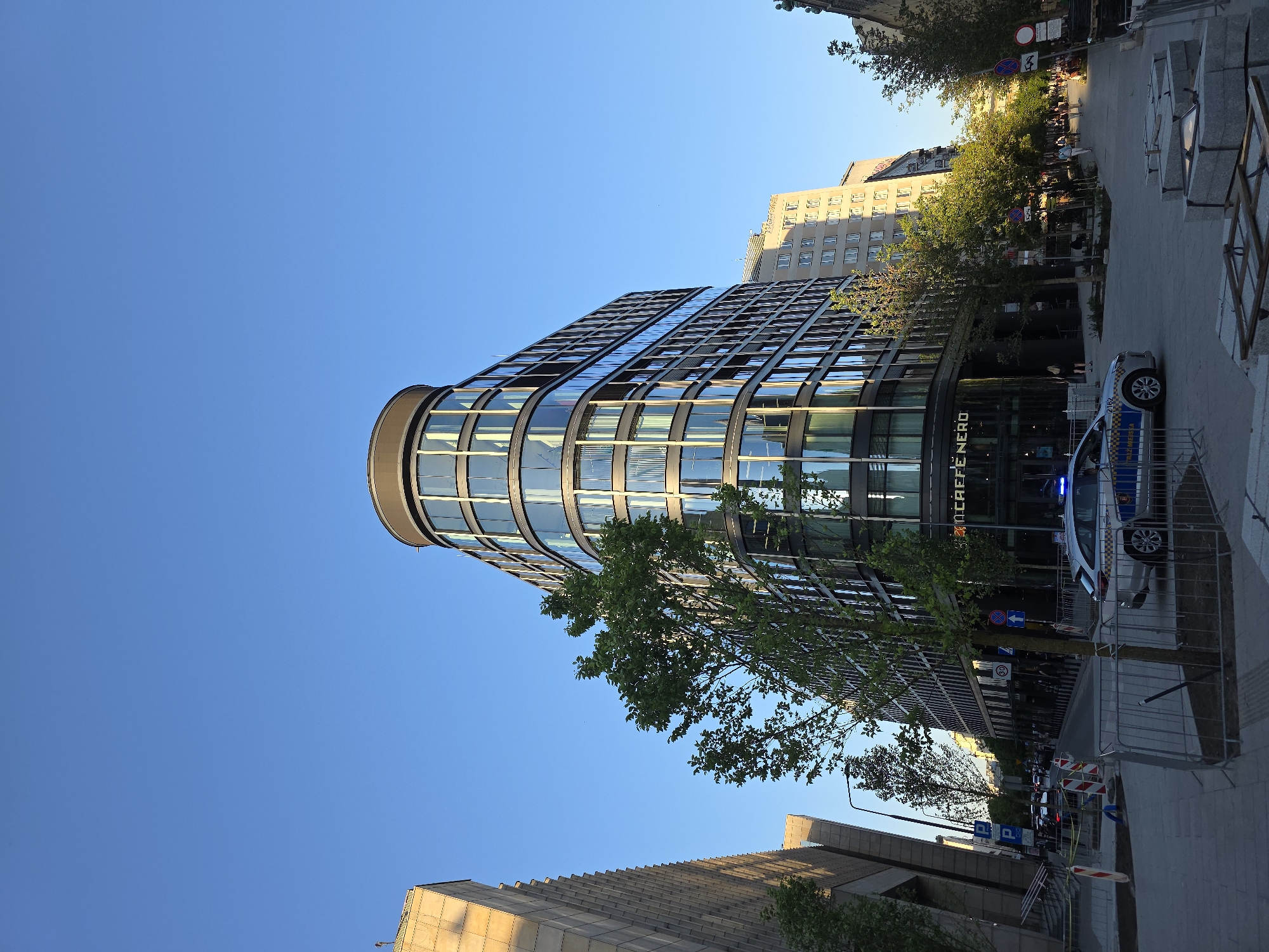 The image shows a street scene in Warsaw, Poland, featuring a modern, multi-story building with a curved glass facade. The building houses a "CAFFE NERO" on the ground floor. A police vehicle with flashing blue lights is parked on the street next to the building, which is partially blocked off with temporary metal barriers. Pedestrians can be seen walking in the background, and trees line the street. A taller building is visible behind the modern structure, while a lower building appears in the foreground. The sky is clear and blue.
