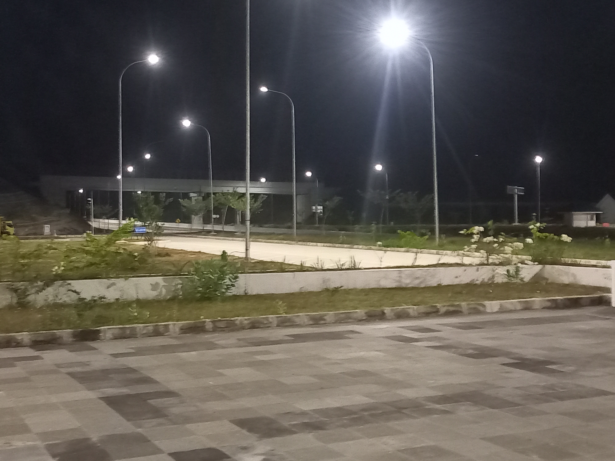 The image depicts an outdoor scene at night, likely a public space or parking lot in Keureuweungkrueng, Indonesia. Several tall streetlights illuminate the area, casting bright light and creating visible lens flare from the camera's perspective. In the foreground, a paved surface with a checkered pattern of gray and darker gray tiles is visible. Beyond this, there is a raised area with sparse green vegetation and concrete borders. Further back, a long, low-lying building structure with illuminated windows is partially visible under the overpass, with several trees and some smaller plants scattered around. The sky is dark, indicating nighttime. The overall impression is one of an open, well-lit area at night, possibly a rest stop or a commercial area. No people or vehicles are clearly identifiable in the image.