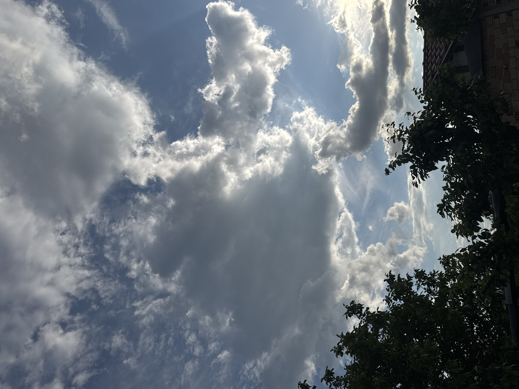 The image shows a partly cloudy sky. Large, white and gray cumulus clouds dominate the frame, obscuring parts of the blue sky. Sunlight filters through gaps in the clouds. In the lower right corner, there is partial foliage, likely from a tree. In the upper right corner is the edge of a building with a brick facade. The location is Sarnevo, Bulgaria.