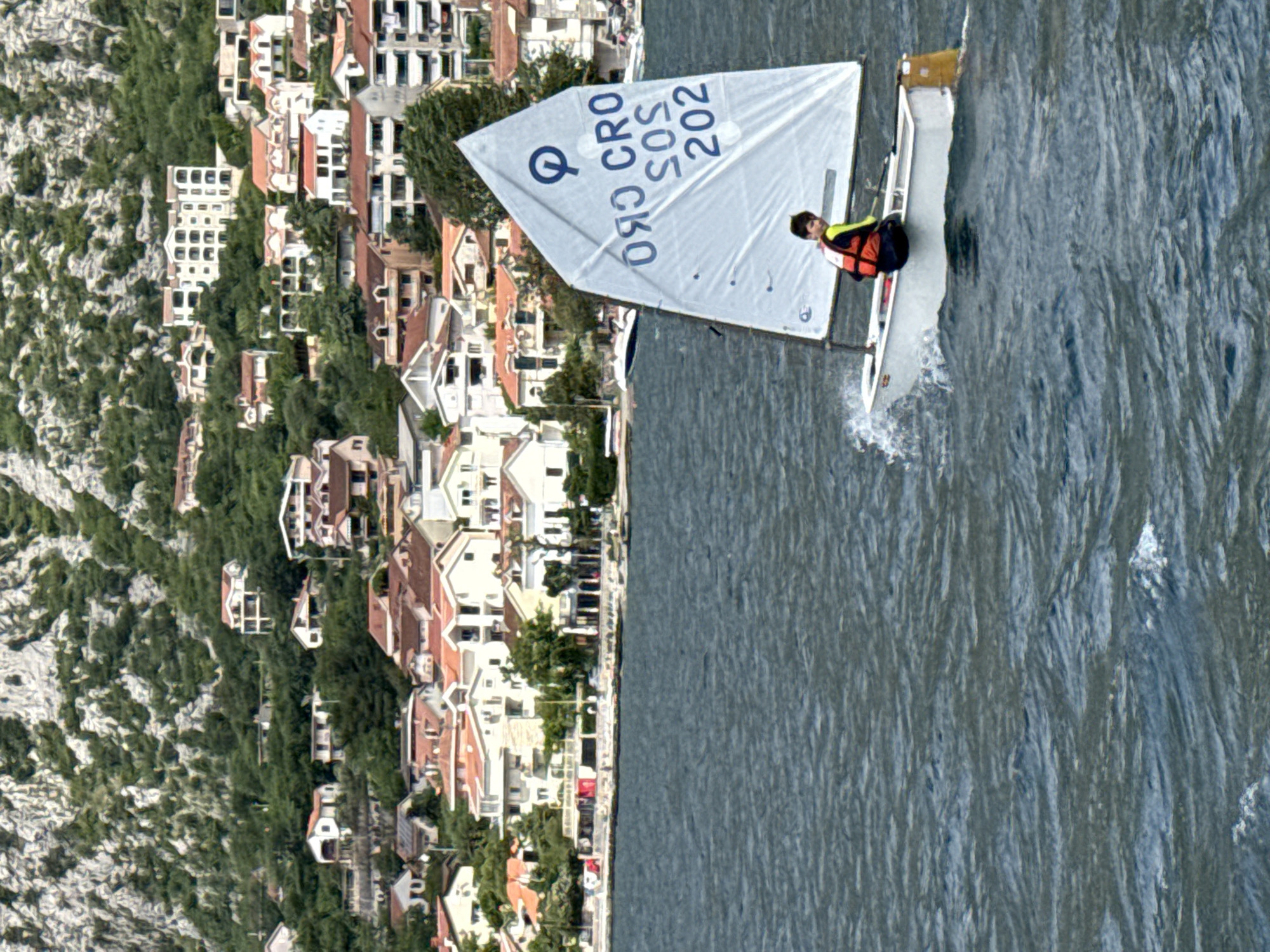 A medium, eye-level shot depicts a lone sailor sailing a small boat on the Bay of Kotor, near Dobrota, Montenegro. The sailboat has a white sail with "Q CRO SOS 202" printed on it. The sailor is wearing a neon yellow jacket and an orange life vest. The waters of the bay are choppy and grey. Along the coast to the left, there is a mixture of buildings and trees. The buildings are a mix of white and red-roofed structures, typical of the region. Part of the mountains is also visible to the left of the buildings.