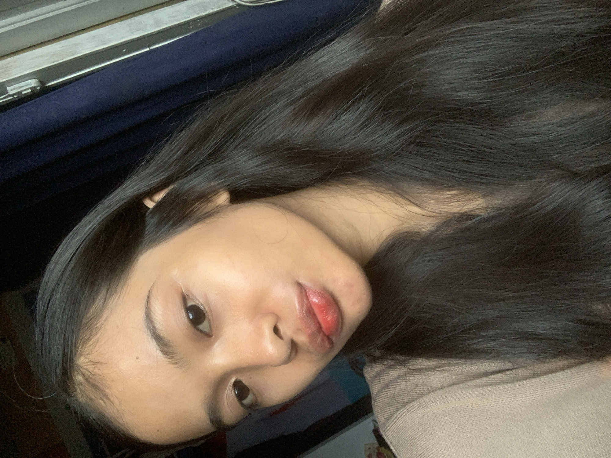 A close-up, head-and-shoulders selfie of a young woman with long, dark hair is taken indoors. She is wearing a beige knitted top. The background includes a dark blue curtain and a partially visible window frame, suggesting an indoor setting. The lighting appears to be natural, coming from the right side of the frame, casting subtle shadows on her face. Her skin is smooth and appears to have a dewy finish, with a hint of blush and lip color. She is looking directly at the camera with a slightly parted lips. No specific geographical markers for Sungai Meriam, Indonesia, are visible in the image.