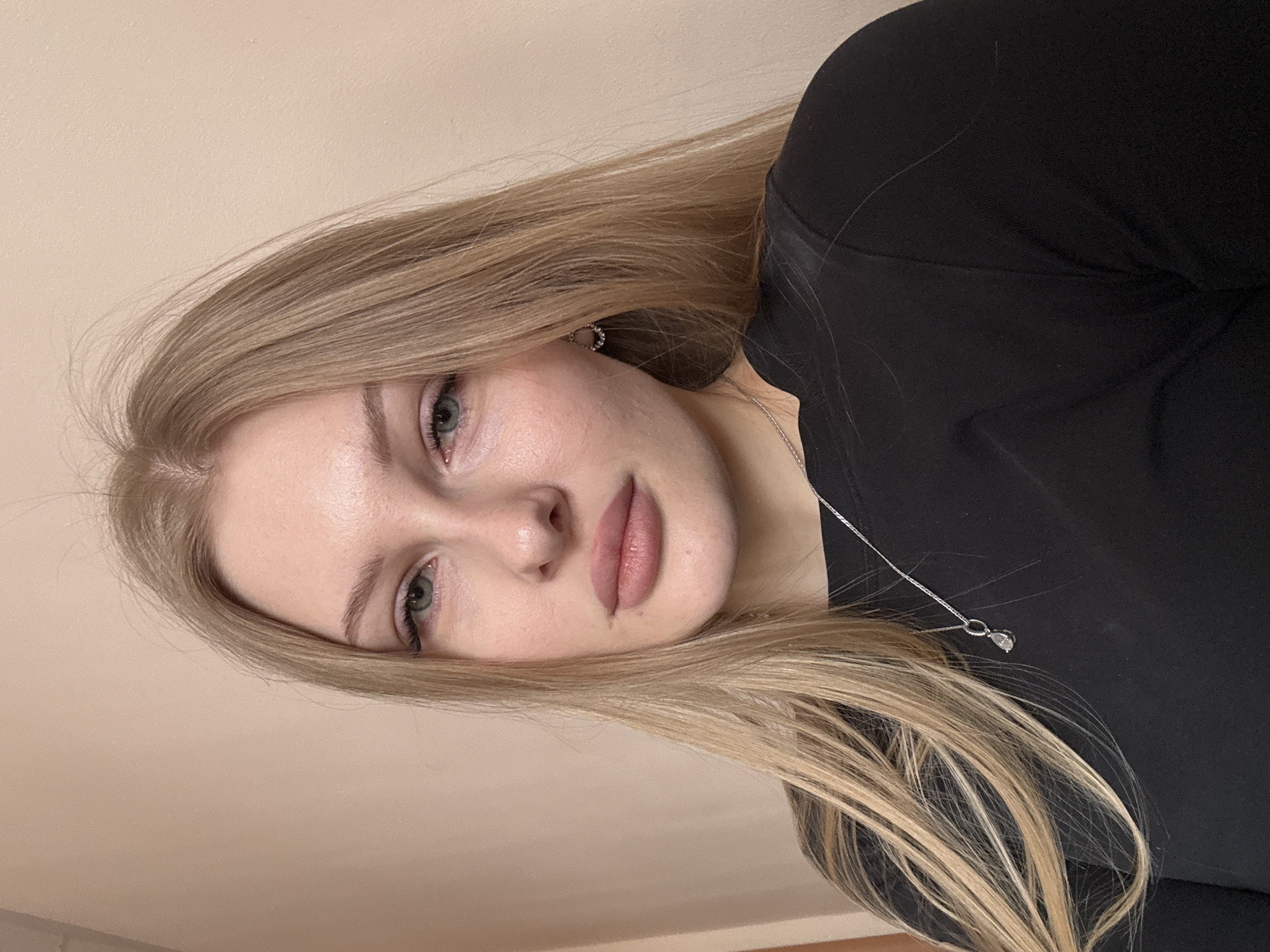 The image depicts a young adult female from the chest up, positioned against a plain, light beige wall. She has long, straight, light blonde hair parted on her left, light-colored eyes, and prominent lips, looking directly at the viewer with a neutral expression. She wears a black long-sleeved top. A silver-colored chain necklace with a clear tear-drop shaped pendant rests on her chest, and a small silver-toned hoop earring with clear stones is visible on her right ear. The indoor setting provides no specific visual cues to determine a location within Perm, Russia.