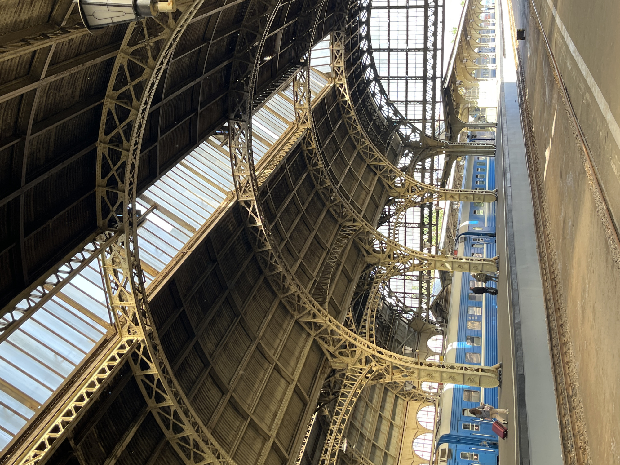 This is a vertical, eye-level shot of the interior of a train station with a blue train on a platform. The station features a large, arched, metal and glass roof structure with visible beams and supports. The train is on a platform to the left, with a few people visible. One person, a woman with a red suitcase, is walking away from the camera on the platform. Another group of three people are standing further down the platform near the train. The train is blue with white stripes and has multiple windows. Tracks are in the foreground. The overall lighting is bright, suggesting daytime. The architectural style of the station appears to be historical. Based on the architecture and the blue and white trains, this is likely Moskovsky railway station in Saint Petersburg, Russia, a historic station known for its grand iron and glass roof.