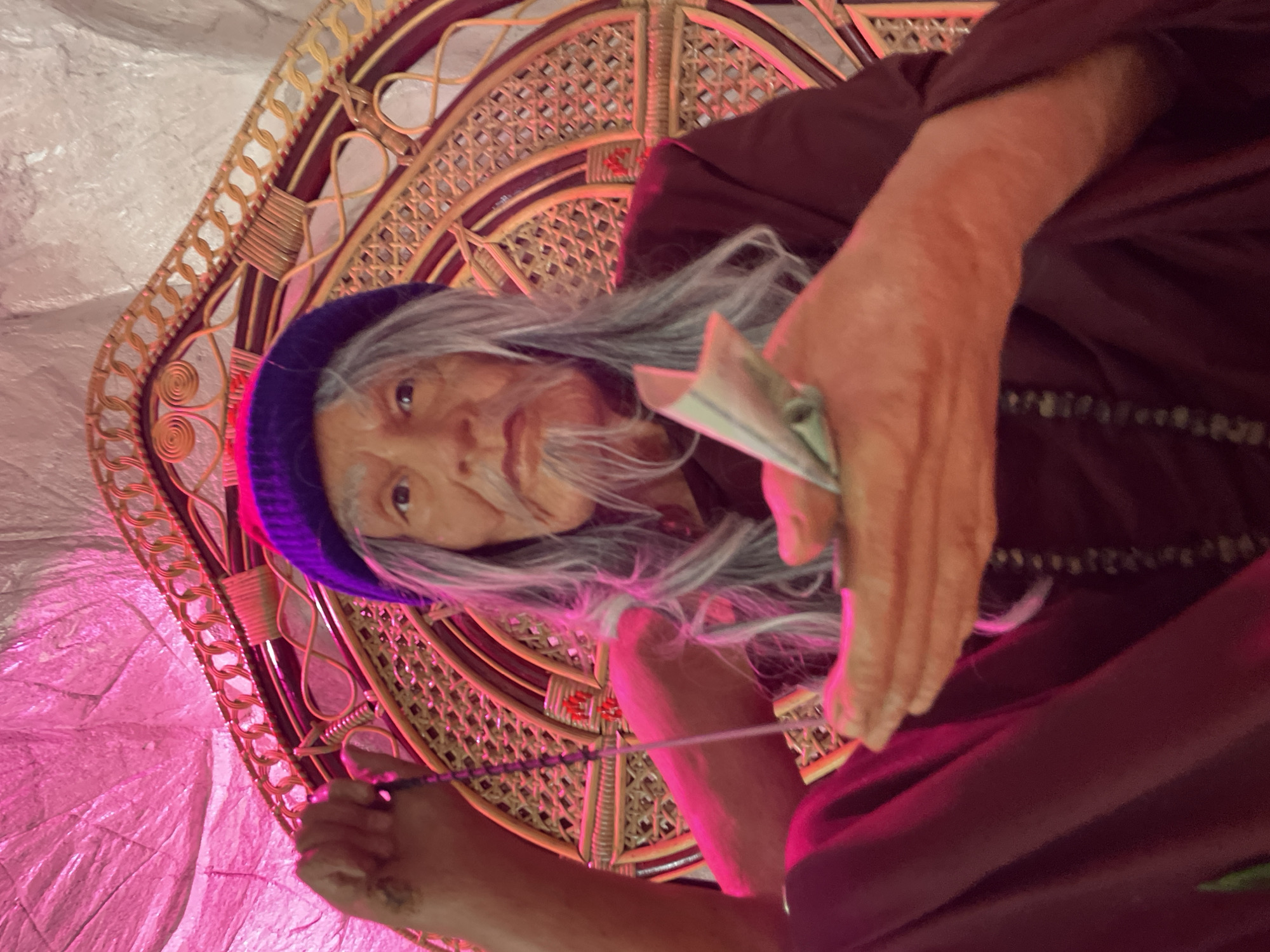 A wax figure of an elderly man with gray hair and a beard, wearing a purple knit cap, a maroon robe, and a rosary, sits in a decorative rattan chair. The figure holds a string of prayer beads in its left hand and a wad of folded bills in its right hand. The background is a textured pink surface and a white wall. The scene is illuminated by pink and red lighting. The image's metadata does not contain information about the city of Bo Win Subdistrict, Thailand.