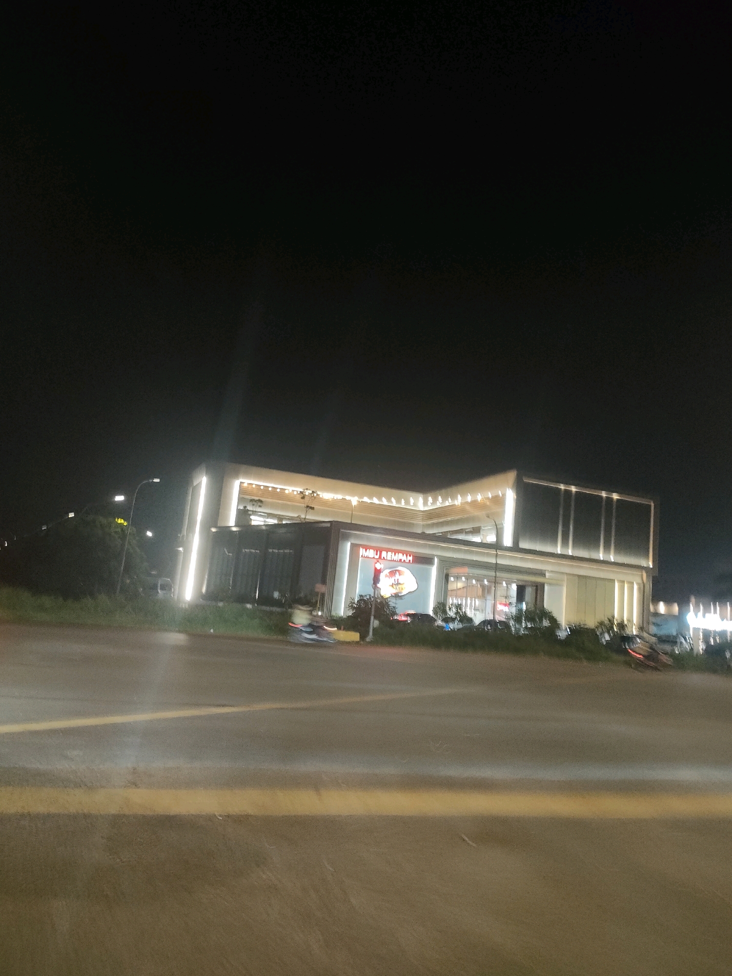 The image depicts a modern, brightly lit building at night, likely a commercial establishment or restaurant, located in Batam, Indonesia. The building features a sleek, multi-level design with illuminated accents and large windows. The facade displays prominent signage, one of which reads "IMBU REMPAH". Several vehicles, including cars and motorcycles, are visible parked or driving in front of the building, suggesting it is a busy location. The surrounding area appears to be a roadside with some greenery and streetlights. The road in the foreground has yellow lane markings. The sky is dark, indicating it is nighttime.