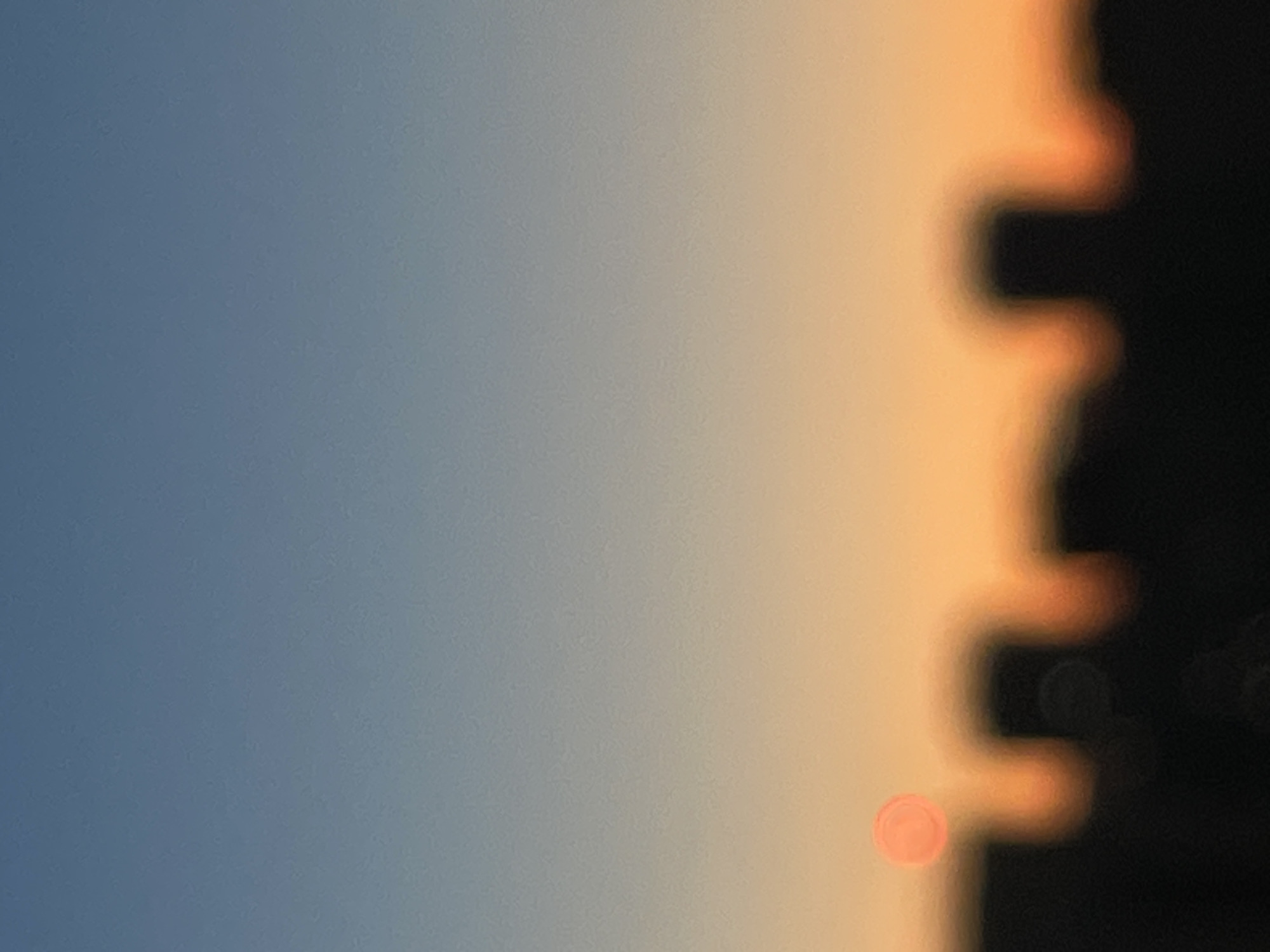 The image depicts a blurred view of a city skyline against a gradient of sky colors, transitioning from a pale blue at the top to an orange-yellow near the horizon. A distinct, out-of-focus red circular object, likely the sun, is visible just above the city silhouette. The skyline itself is rendered as dark, indistinct shapes, suggesting buildings. The overall impression is of a sunset or sunrise over an urban area. The location in Sinyavino, Russia, is noted, but there are no specific visual cues in the image to confirm this location, as it is a general cityscape without identifying landmarks. There are no people, animals, or specific objects identifiable within the frame due to the blurriness.