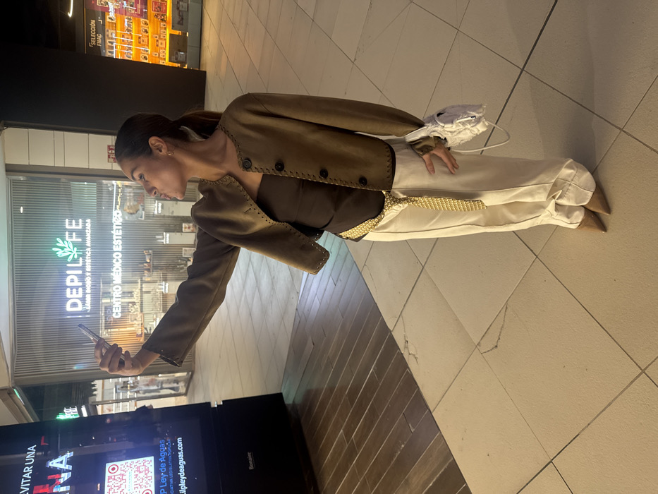 An image taken inside a commercial shopping center, specifically the BonAire shopping center in Aldaia, Spain. A female subject, of slender build and medium complexion, is positioned slightly off-center to the right, engaged in taking a selfie with a silver-colored smartphone held aloft in her right hand. Her head is tilted to the left, and her lips are pursed. She wears a dark brown strapless top, a short brown jacket with decorative stitching and four large dark buttons, wide-leg cream trousers, a long gold chain belt draped across her waist, and pointed-toe brown shoes. A crumpled white bag with a strap is held in her left hand.
The background features multiple commercial establishments. To the immediate left is a tall, vertical digital display screen exhibiting text in Spanish, including "EVITAR UNA" (to avoid a), a QR code, "LEY DE AGUAS" (Water Law), and "BonAire" at the bottom. In the center background, a storefront with a glass facade displays "DEPI LIFE" in prominent green and white lettering, accompanied by "JAVIER MONXO Y ESTETICA AVANZADA" and "CENTRO MÉDICO ESTÉTICO" (Medical Aesthetic Center). Merchandise is visible through the glass. To the far right, another storefront is partially visible, showing "SELECCIÓN FNAC" (FNAC Selection). The floor consists of light beige square tiles with visible cracks in several places, transitioning to a section of dark wood-patterned flooring on the far left.