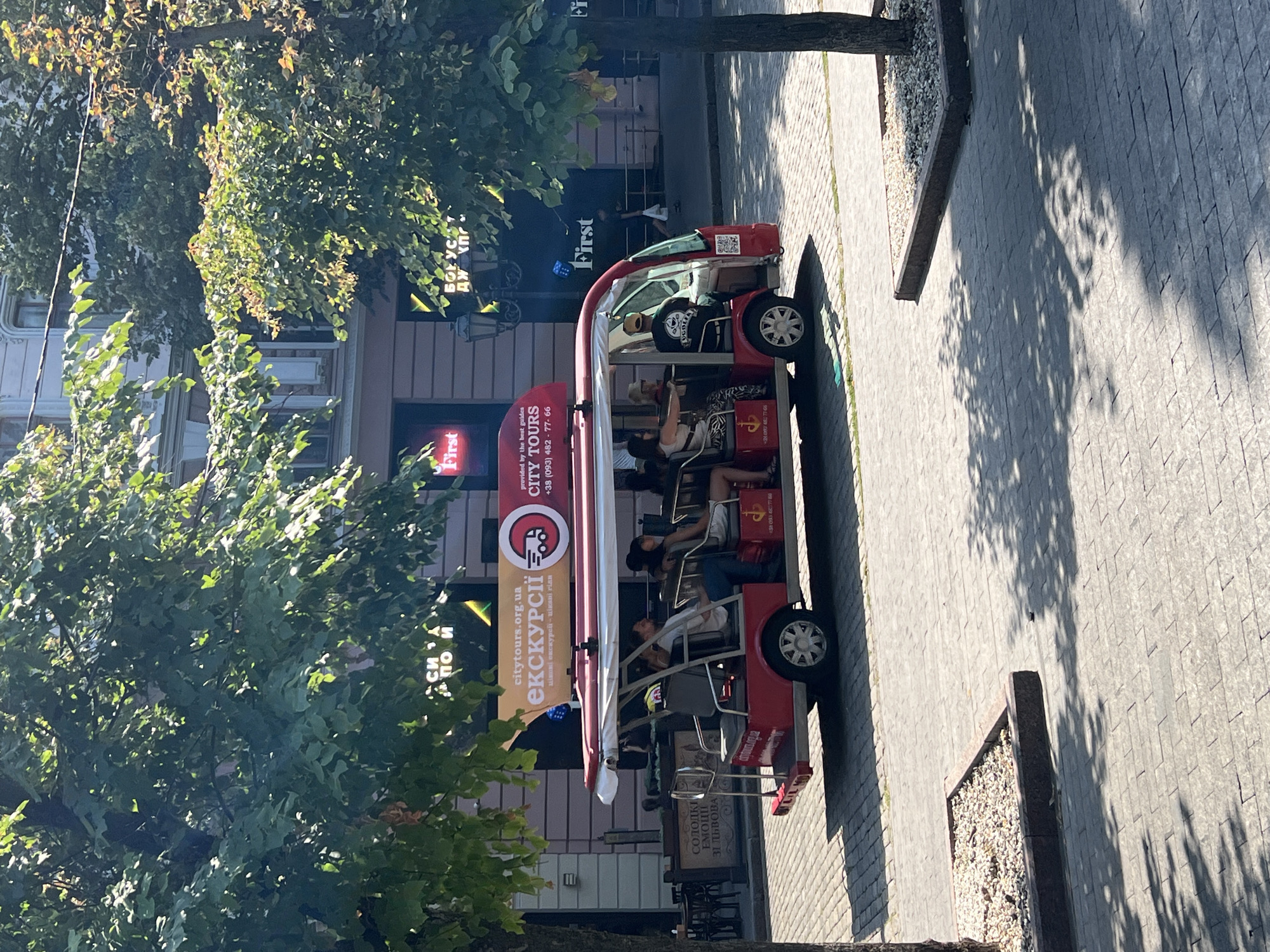 A red, open-air tour vehicle with a canopy is parked on a cobblestone street. The vehicle has seats for passengers, and several people are seated inside. A banner above the vehicle advertises "citytours.org.ua" and "CITY TOURS". The scene appears to be in a city, with buildings and trees visible in the background. The overall impression is that of a tourist activity in a European city.