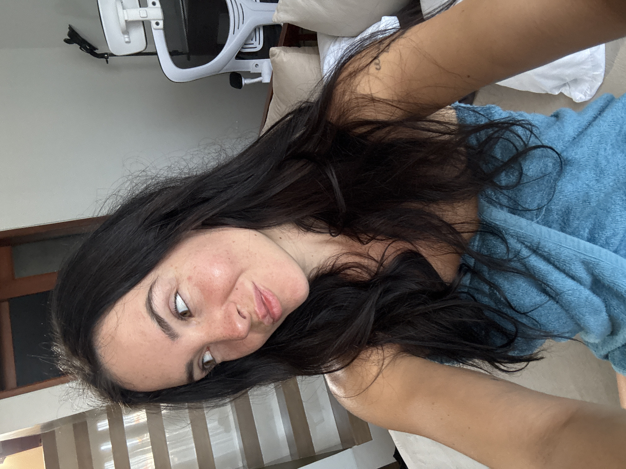 A young woman with long, dark hair and freckles is taking a selfie in a room in North Kuta, Indonesia. She is wearing a blue towel wrapped around her chest. She is looking away from the camera with her lips slightly pursed. In the background, a white office chair with a black headrest is visible, along with a set of stairs and wooden doors. There are also pillows on a bed or couch.