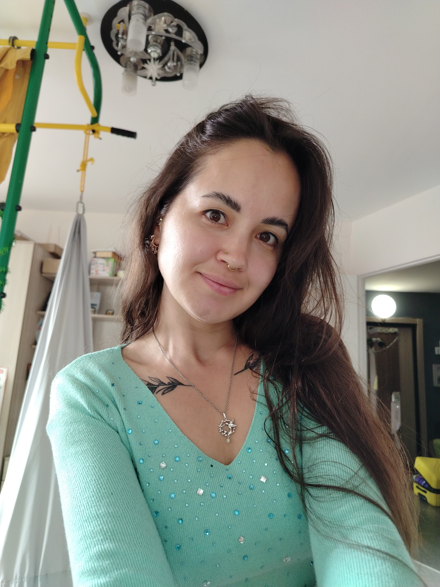 A young woman with long, dark hair and brown eyes is pictured indoors. She is wearing a light teal, long-sleeved, V-neck top adorned with small, iridescent blue and clear embellishments. A silver necklace with a circular pendant featuring a bird and floral motif rests on her chest. She has a tattoo of a branch with leaves on her sternum and a nose ring. Her left ear displays a gold stud earring. She is looking directly at the camera with a slight smile.
The background reveals an interior setting. To the left, a green and yellow gymnastic apparatus is visible, with part of a gray hammock hanging from it. Behind her, shelves with various items, including boxes and what appear to be books, are present. A white, gauzy fabric hangs on the left side of the frame. To the right, a doorway leads to another room with a round light fixture visible. The overall environment suggests a home or recreational space. The location in Noviy, Russia, is not discernible from the visual information in the image.