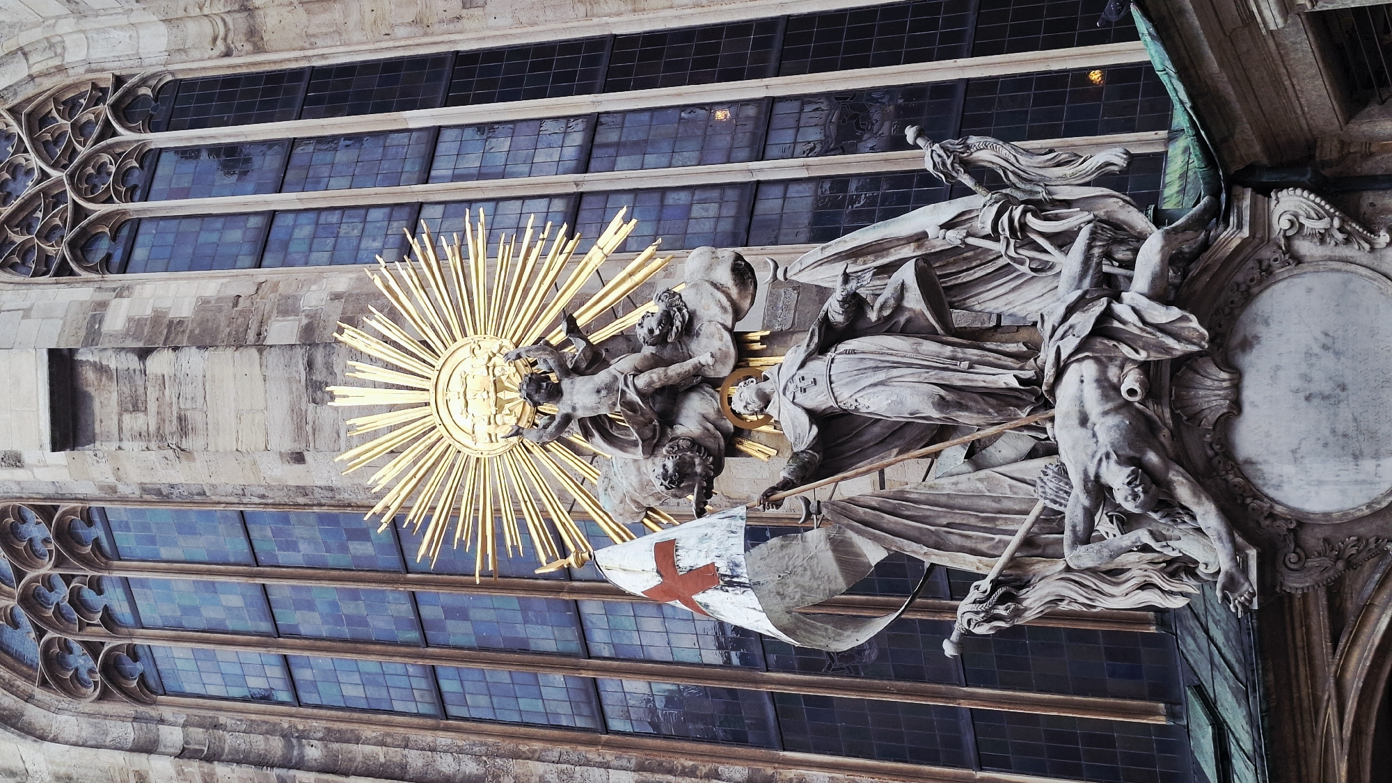 The image shows a section of the Stephansdom (St. Stephen's Cathedral) in Vienna, Austria, specifically the High Altar area. The central focus is a baroque sculpture, likely part of the altar, featuring several figures. A golden sunburst emanates from behind some of the figures, drawing attention. One figure seems to be a cleric, holding a flag with a red cross on it. Another figure appears to be an upside-down man. The backdrop is comprised of Gothic-style windows with leaded glass, dividing the light into a grid-like pattern. The architectural style of the cathedral is clearly visible in the stonework and detailing surrounding the sculpture.