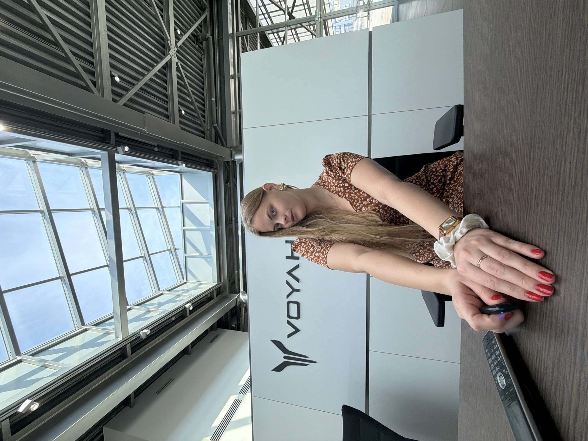A woman with long blonde hair sits at a dark wooden desk in a modern office setting. She wears a floral print dress and a white scrunchie on her wrist. Her nails are painted red. She is holding a small black device in her hands, and a remote control is on the desk. Behind her is a white wall with the logo "VOYAH" displayed. Large windows with a metal framework offer a view of the outside. The location is Izhevsk, Russia.