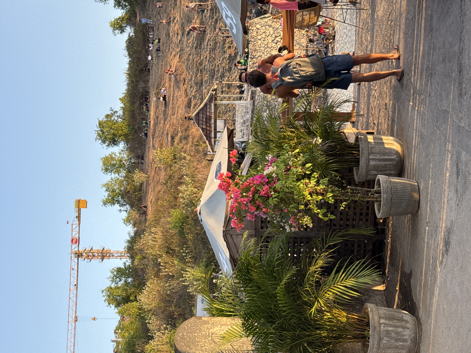 In Kuta Selatan, Indonesia, a construction crane is visible on a hillside. Numerous people are scattered across the grassy incline, some sitting and others standing, observing the scene. In the foreground, a man with a backpack faces away from the camera, looking towards a sandy area where other individuals are present. Two large potted plants, one a palm and the other with pink bougainvillea, frame the immediate foreground. Several structures with tent-like roofs are present, suggesting an outdoor gathering or seating area. The lighting indicates it is late afternoon, with long shadows cast across the concrete ground.