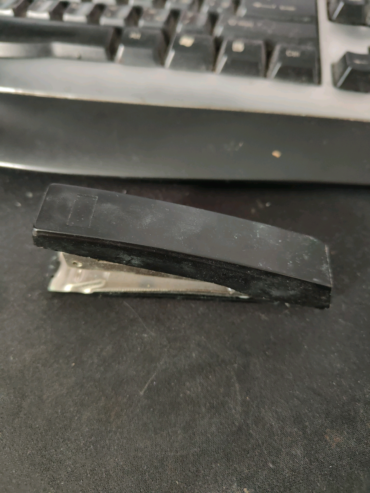 A black stapler rests on a textured dark surface, possibly a desk, in front of a keyboard. The stapler is closed, with its metal base slightly extended from the black plastic top. The keyboard appears to be a standard computer keyboard, with gray keys and black lettering. The environment suggests an indoor setting, likely a workspace. The specific location in Homyel, Belarus, cannot be determined from the image alone.