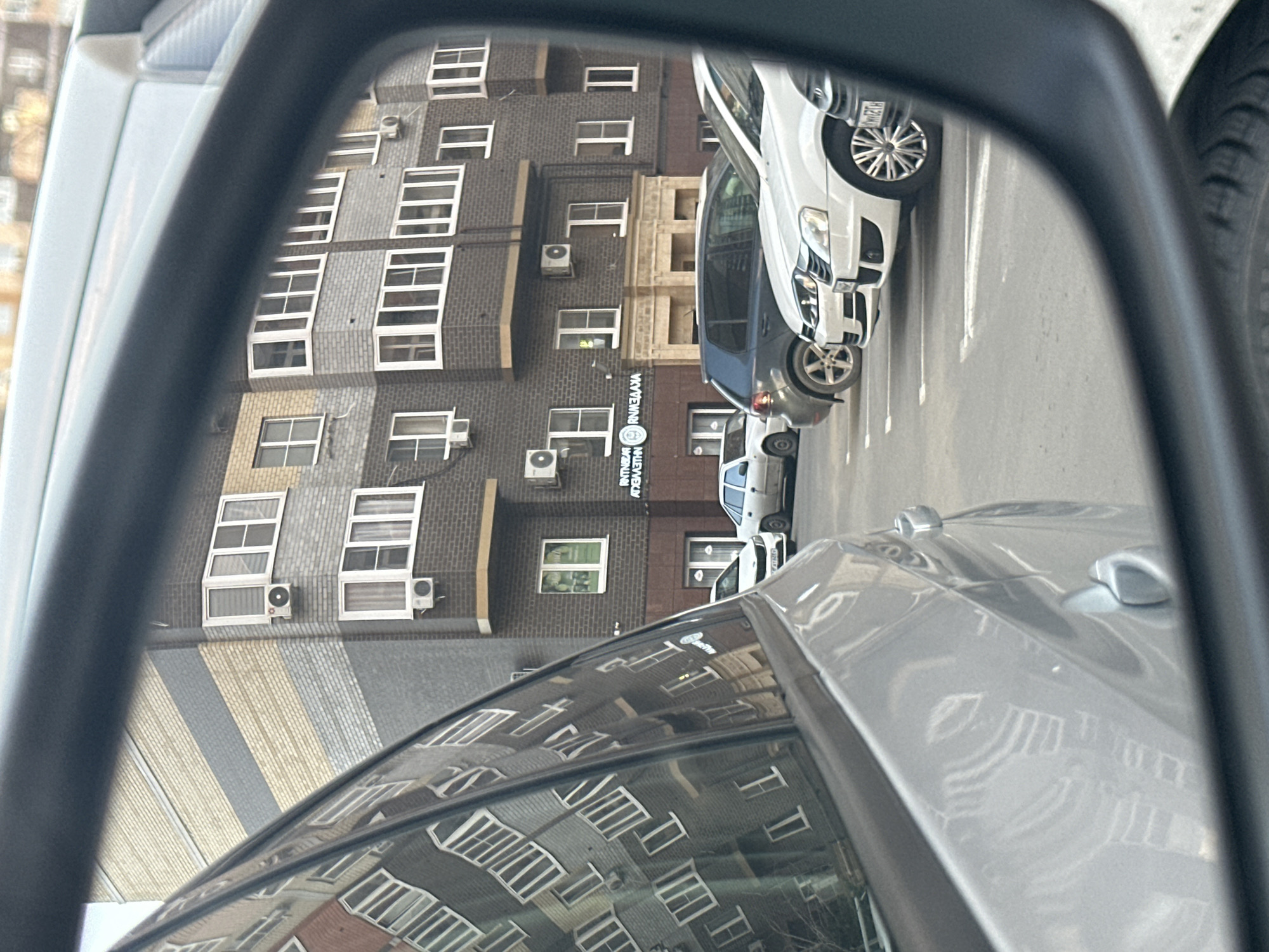 The image presents a street scene in Rostov-on-Don, Russia, observed through the passenger-side mirror of a vehicle. The specific location is identified as the area around Kazakhskaya Street, 93A.
The primary subject visible within the mirror is a multi-story building constructed with alternating dark grey, light grey, and beige brick facades. This building features numerous white-framed windows, several of which are bay windows, and multiple external air conditioning units affixed to its exterior. The ground level includes a commercial unit displaying a circular sign and illuminated Cyrillic text, reading "ЯНТНЕВЪ" and "ЯНАВЛАХА," situated above a beige stone-clad entrance.
In front of this building, a paved parking area with visible white painted lines contains several parked vehicles. From foreground to background within the mirror's view, these include a white sedan with distinctive multi-spoke hubcaps and a partial license plate displaying "M H22C H," a dark grey hatchback, and a lighter-colored sedan.
The immediate foreground of the image shows the silver bodywork and door handle of the vehicle from which the photograph was taken, along with the dark frame of the side mirror. A reflection of another brick building, primarily reddish-brown in hue, is visible on the side window of the originating vehicle. No individuals are discernible in the scene.
