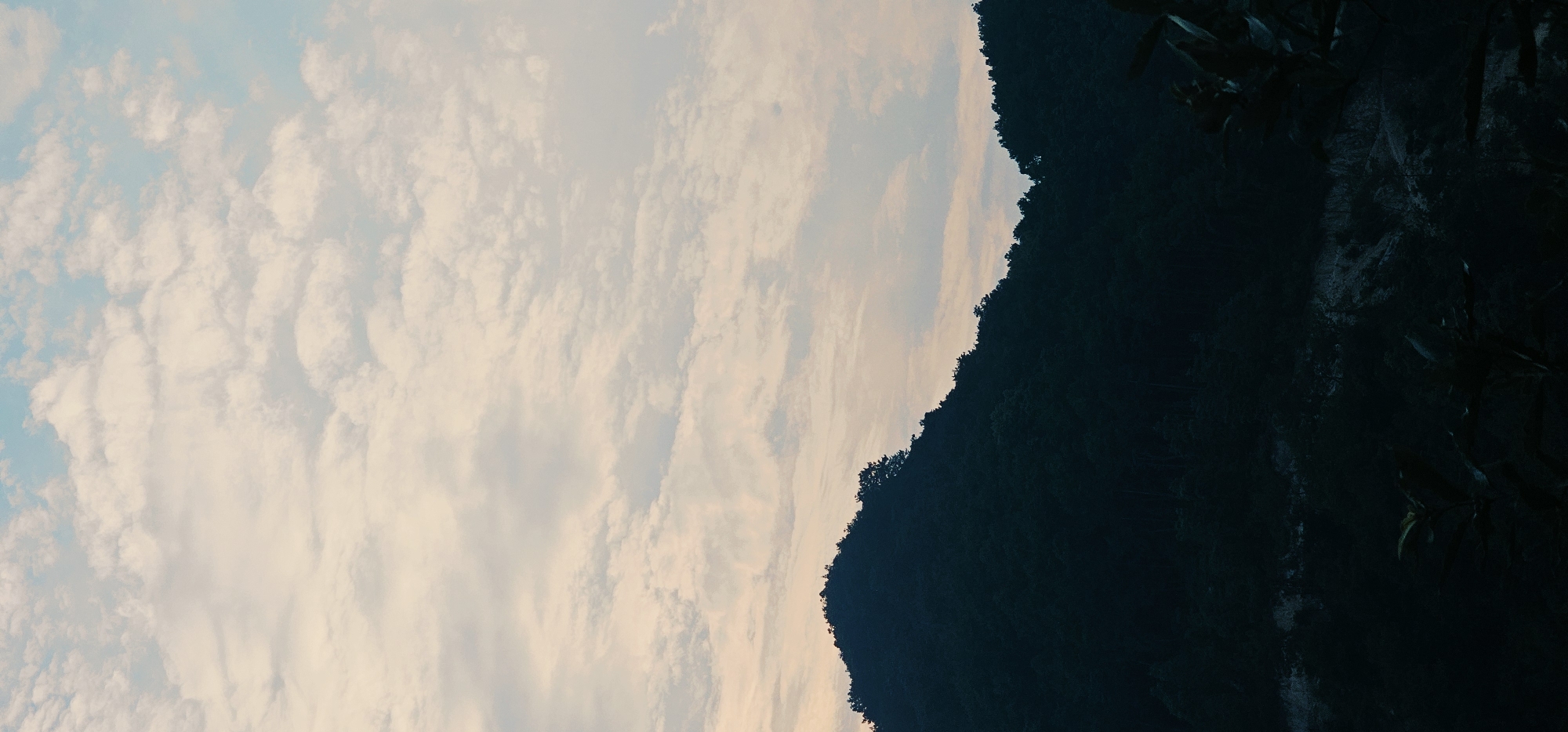 A dark, forested mountain range dominates the lower half of the image, silhouetted against a pale, cloud-filled sky. The clouds are a mix of white and light pink, suggesting either sunrise or sunset. The mountain slopes are densely covered with trees, and the foreground shows a dark, rocky embankment with some sparse vegetation. The scene appears to be a natural landscape, likely in a mountainous region. The specific location in the Chongqing High-tech Zone, China, is not visually identifiable from the image itself, as it depicts a natural environment devoid of any urban or technological markers.