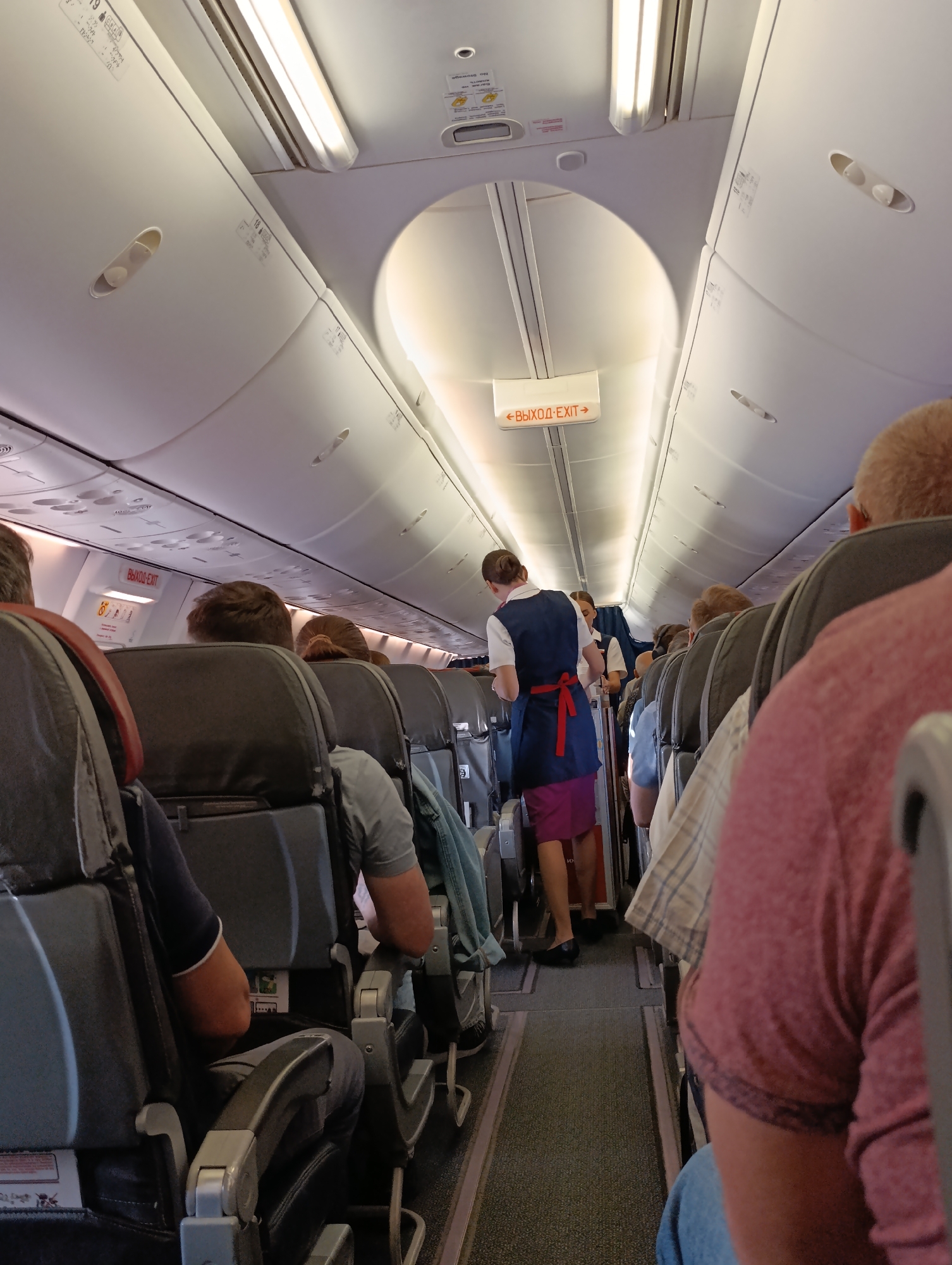 The image captures the interior of an airplane cabin, showing rows of passenger seats and an aisle. Several passengers are seated, facing forward. In the aisle, two flight attendants are visible. One, wearing a blue vest over a white shirt and a purple skirt, appears to be attending to passengers. Another flight attendant is partially visible behind her. An illuminated exit sign, reading "ВЫХОД-EXIT," is mounted on the ceiling. The lighting in the cabin is from overhead fluorescent fixtures. The scene suggests a typical flight environment with passengers and crew in their designated areas. The image does not provide any specific visual cues that would indicate the location in the city of Staroe Mart'yanovo, Russia.