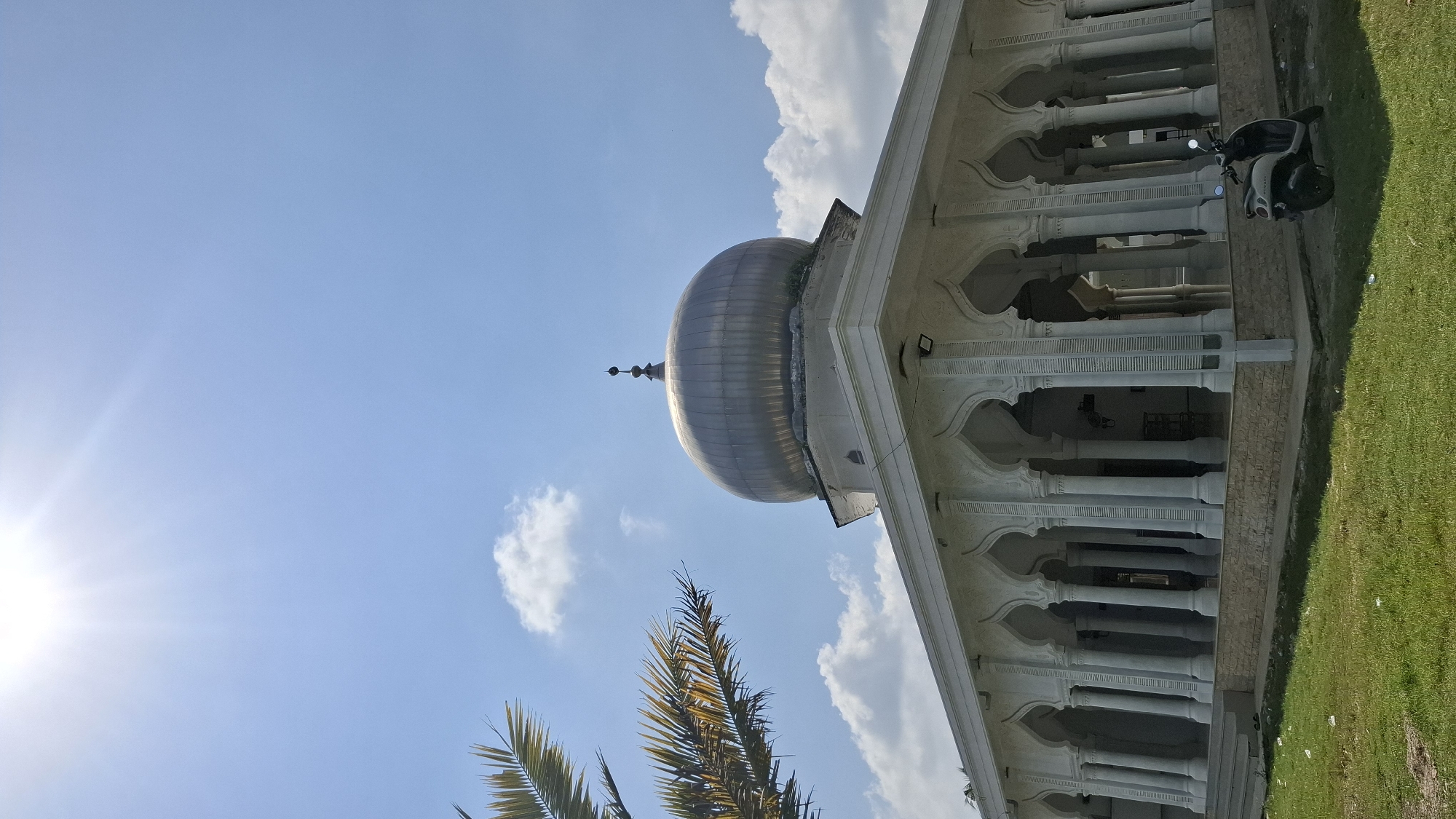 In Grong-grong, Indonesia, a bright, sunny day illuminates a mosque with white walls and a prominent silver dome. The mosque features an arcade of white, arched colonnades supporting the roof. In the foreground, a lush green lawn is visible, with a few scattered pieces of white debris. A white scooter is parked on the grass to the right of the mosque. Palm fronds from a tree frame the upper left corner of the image against a clear blue sky dotted with white, fluffy clouds. The sun is high and bright, casting a strong light.