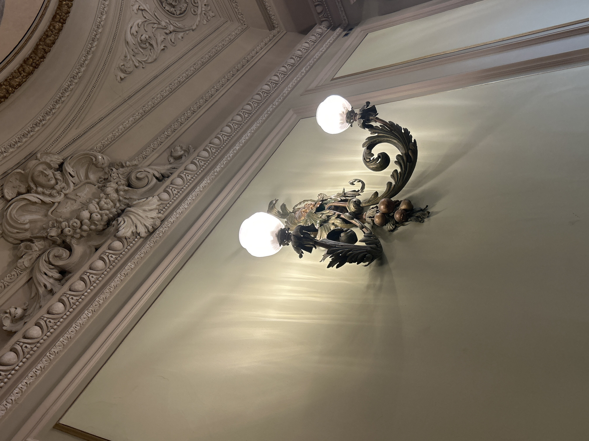 This upward-angled photograph captures a richly decorated interior, characteristic of grand historic architecture, consistent with a setting in Milan, Italy. The scene features an ornate, dark-toned metal wall sconce with two glowing, frosted spherical light bulbs. The sconce itself is elaborately designed with intricate scrollwork, possibly depicting stylized leaves, small fruits (such as berries or grapes), and a subtle bird-like element. The light emitted from the bulbs creates distinct, wavy patterns of illumination and shadows on the light-colored wall.
To the left and extending across the upper part of the frame, the ceiling and upper wall are adorned with multiple layers of elaborate stucco or plaster molding. These decorative elements include detailed cornices and friezes featuring classical motifs such as continuous scrolls, bead patterns, and intricate floral designs. A prominent sculptural relief in the lower-left section depicts a classical figure, resembling an angel or cherub with wings, surrounded by fruit and foliage. A textured gold-colored trim runs along the uppermost edge of the ceiling's decorative elements.
The color palette is predominantly off-white, cream, and light beige for the architectural details, providing a stately backdrop for the dark, metallic sconce and the warm, artificial light it casts. The perspective highlights the height and detailed craftsmanship of the interior. There is no visible activity or text. The dominant artificial lighting suggests an indoor setting, likely in the evening or a dimly lit space.