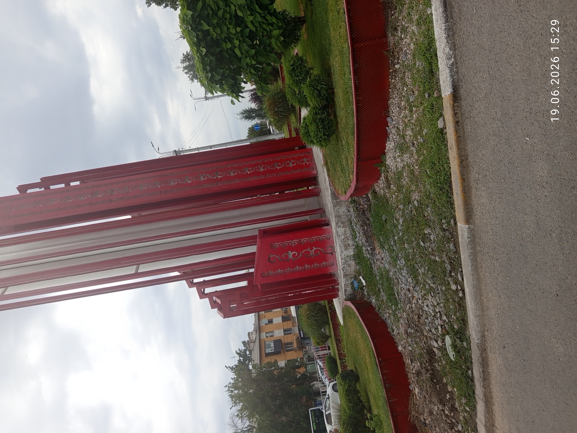 The image shows a monument in Bishkek, Kyrgyzstan. The monument is made of red and white vertical elements, with decorative patterns on the red sections. In the background, there are buildings, trees, streetlights, and a bus. The scene appears to be an urban setting on a cloudy day. The timestamp "19.06.2026 15:29" is visible in the lower right corner of the image.