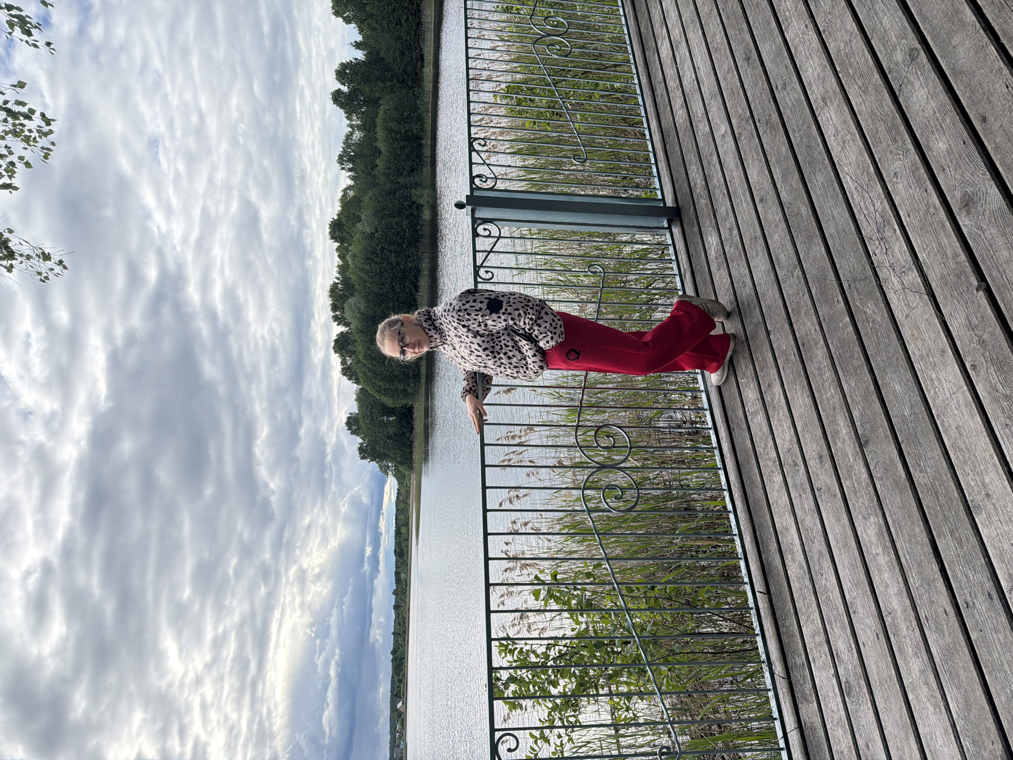 A woman is standing on a wooden deck overlooking a body of water in Zaplav'e, Russia. She is wearing a spotted jacket and red pants. A black metal railing separates the deck from the water. Vegetation grows along the water's edge. The sky is cloudy.