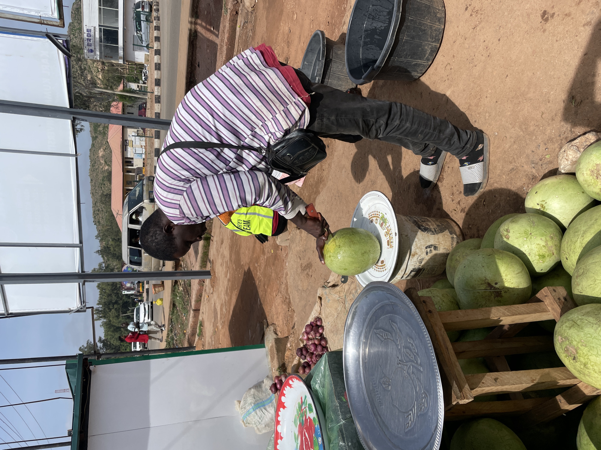 An outdoor scene depicts a man, likely a vendor, attending to his produce at a roadside stall in what appears to be Gura Top, Nigeria. The man, with short dark hair, is wearing a purple and white striped short-sleeved shirt over a red undershirt, black trousers, and patterned socks with white and black slides. A small black shoulder bag, with "BOSS" visibly printed on it, is slung across him, and a yellow safety vest with "URLEY CLEAN" partially visible is worn underneath his shirt. He is holding a small, light green watermelon in one hand and a small knife in the other, possibly preparing to cut or inspect the fruit.
Beside him, an assortment of watermelons, also light green, are stacked in a wooden crate and scattered on the dusty ground. Nearby, a small pile of red onions sits on the ground. Various metallic and patterned trays, including one silver tray with an apple design and another white one with a red floral pattern, are also part of the display. Several black plastic tubs or buckets are seen on the brown, earthy ground.
The scene is brightly lit by strong sunlight, casting distinct shadows, indicating a clear, sunny day. In the background, a paved road stretches with a white van and another white car visible. Beyond the road, buildings with white facades, one appearing to be a "PHARMACY" (partially visible and inverted), are situated against a backdrop of distant hills or mountains. A large white billboard structure and power lines are also present, adding to the urban-rural blend of the setting.