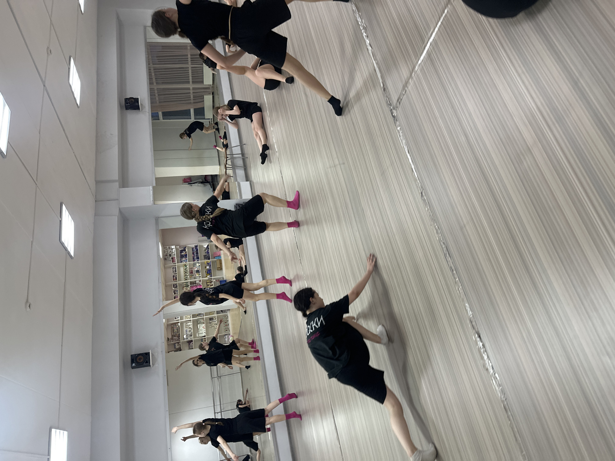 The image shows a group of young dancers in a dance studio, likely located in Votkinsk, Russia, given the lettering on their shirts. The studio has a wooden floor and mirrored wall. The dancers are wearing black outfits, and some have pink socks. They are performing a dance routine, with their arms extended and bodies angled. The studio is brightly lit, and there's a bookshelf visible in the background, indicating it could be a community center or school dance studio.