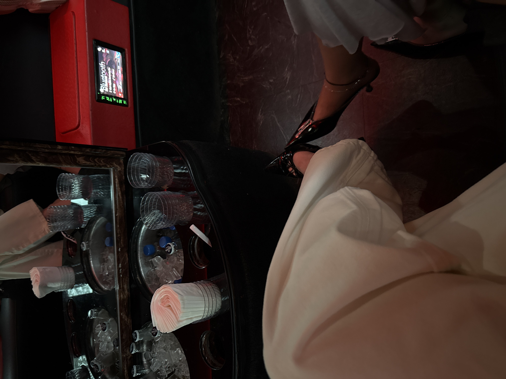 The image shows the interior of a dimly lit space, possibly a nightclub or lounge in Miami Beach, United States of America. A woman in black heels with an ankle bracelet is seated. A red Bluetooth device sits on a bar with cups, napkins, ice, and bottles.