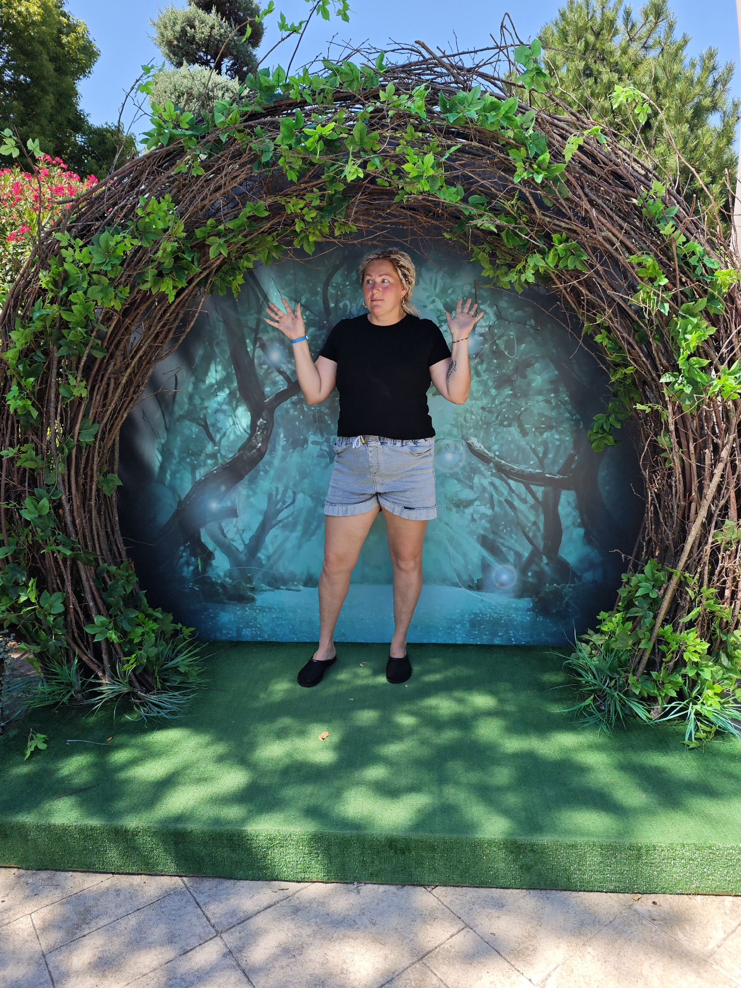 A person stands in the center of a nest-like archway decorated with branches and green leaves, in front of a backdrop depicting a mystical forest. The person, who appears to be female, is wearing a black t-shirt, denim shorts, and black slip-on shoes. Her arms are raised with palms facing outward, and she has blonde hair pulled back. The archway is set on a green carpeted platform. The backdrop is a printed image of a dark, enchanted forest with glowing orbs. The scene is outdoors, with trees and a bright blue sky visible above the archway. The ground in front of the platform is paved with square tiles, and shadows of leaves are cast on the green carpet. This scene appears to be a photo opportunity set up in a public area, possibly a park or an entertainment venue in the city of Sochi, Russia.