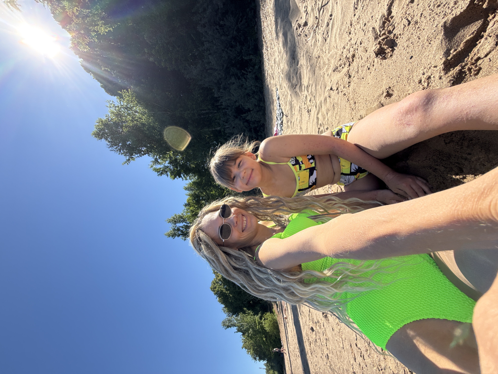 This image, captured in Шебунино, Russia, displays an adult female and a child female seated on a sandy bank under a clear blue sky on a bright, sunny day. The adult, positioned on the left, possesses long, blonde dreadlocks, wears oval-framed sunglasses, and is dressed in a neon green textured one-piece swimsuit. Her right arm is extended, holding the camera for a selfie. The child, seated to the right of the adult, has brown hair with bangs and wears a yellow two-piece swimsuit featuring cartoon character prints; her right knee exhibits visible skin irritation. Both individuals are smiling directly at the camera. The background consists of a dense line of green trees and bushes forming an embankment. Direct sunlight from the upper right corner produces prominent lens flare. Several blurry figures are discernible on the distant sandy area to the far left, and a patterned blanket or towel is partially visible on the sand behind the child.