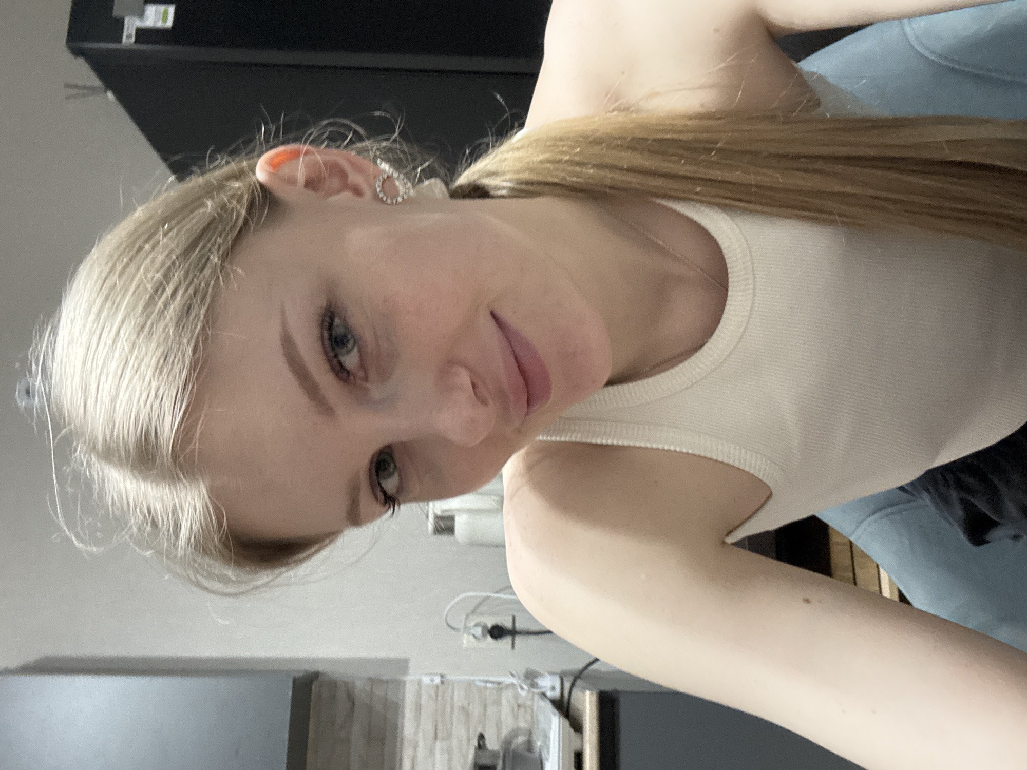 A fair-skinned young woman is captured in a close-up, tilted portrait. Her blonde hair is pulled back into a ponytail, revealing a delicate, teardrop-shaped earring in her left ear. She wears a ribbed, white tank top. The background features a mix of domestic elements: a black refrigerator or cabinet in the upper area, light blue jeans on her lap, a section of a tile backsplash and countertop visible in the lower portion of the frame, along with various indistinct kitchen items. The location is not discernible from the image.