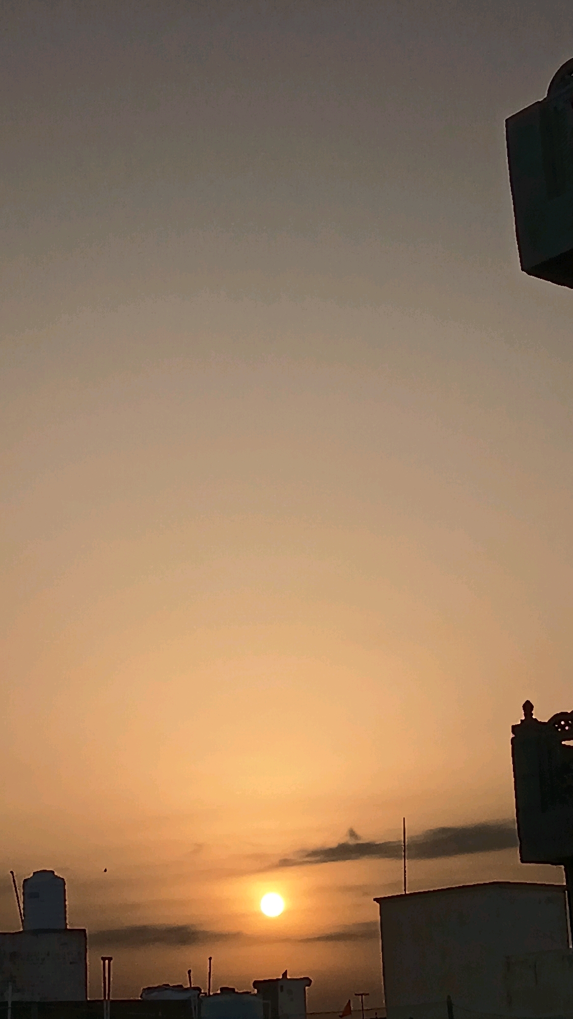 The image captures a sunset over a cityscape. The sky is a gradient of warm oranges and soft browns, with the sun appearing as a bright, diffused orb near the horizon. Silhouetted buildings with flat roofs and some water tanks are visible in the foreground, suggesting an urban environment. A few small, dark clouds are scattered across the sky, adding depth to the scene. The overall impression is one of a peaceful evening in a city.
