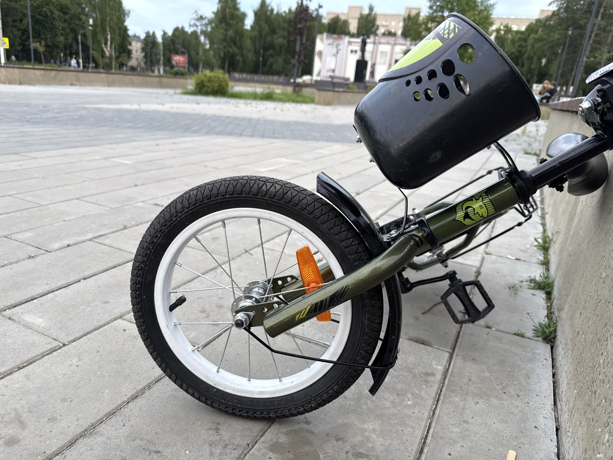 This is a close-up, ground-level shot of a green bicycle parked on a paved outdoor plaza. The bicycle's front wheel, with a white rim and black tire, is prominently featured in the foreground, leaning against a concrete wall. The handlebars and a black basket are visible at the top, and the pedal and part of the frame are also in view. The scene appears to be in Chaikovsky, Russia, with buildings and trees in the background suggesting an urban park or public square. The lighting indicates it might be daytime, possibly overcast. There is no discernible activity or people engaged in actions, suggesting the bicycle is parked. The overall mood is neutral and observational. The tire has some visible text: "14x1.75 (57-203)MAX."
