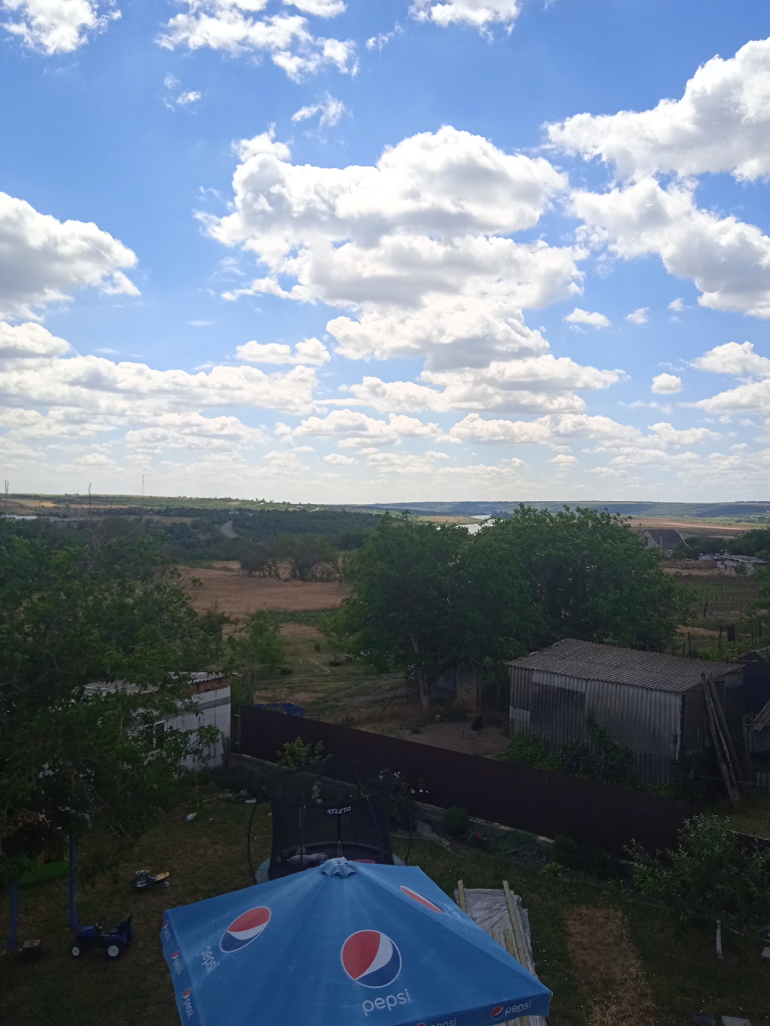 The image shows a rural landscape with a wide expanse of sky filled with white, puffy clouds against a blue background. In the foreground, a blue umbrella with the Pepsi logo is visible, partially covering a trampoline and some lawn furniture. Behind the umbrella, a small, weathered shed with a corrugated metal roof stands next to a fenced-off area. Further in the distance, the landscape transitions into fields and trees, with a body of water visible on the left. In the far distance, the horizon is marked by rolling hills. The scene appears to be located in the village of Rakove, Ukraine, based on the context provided. There are no people visible in the image.