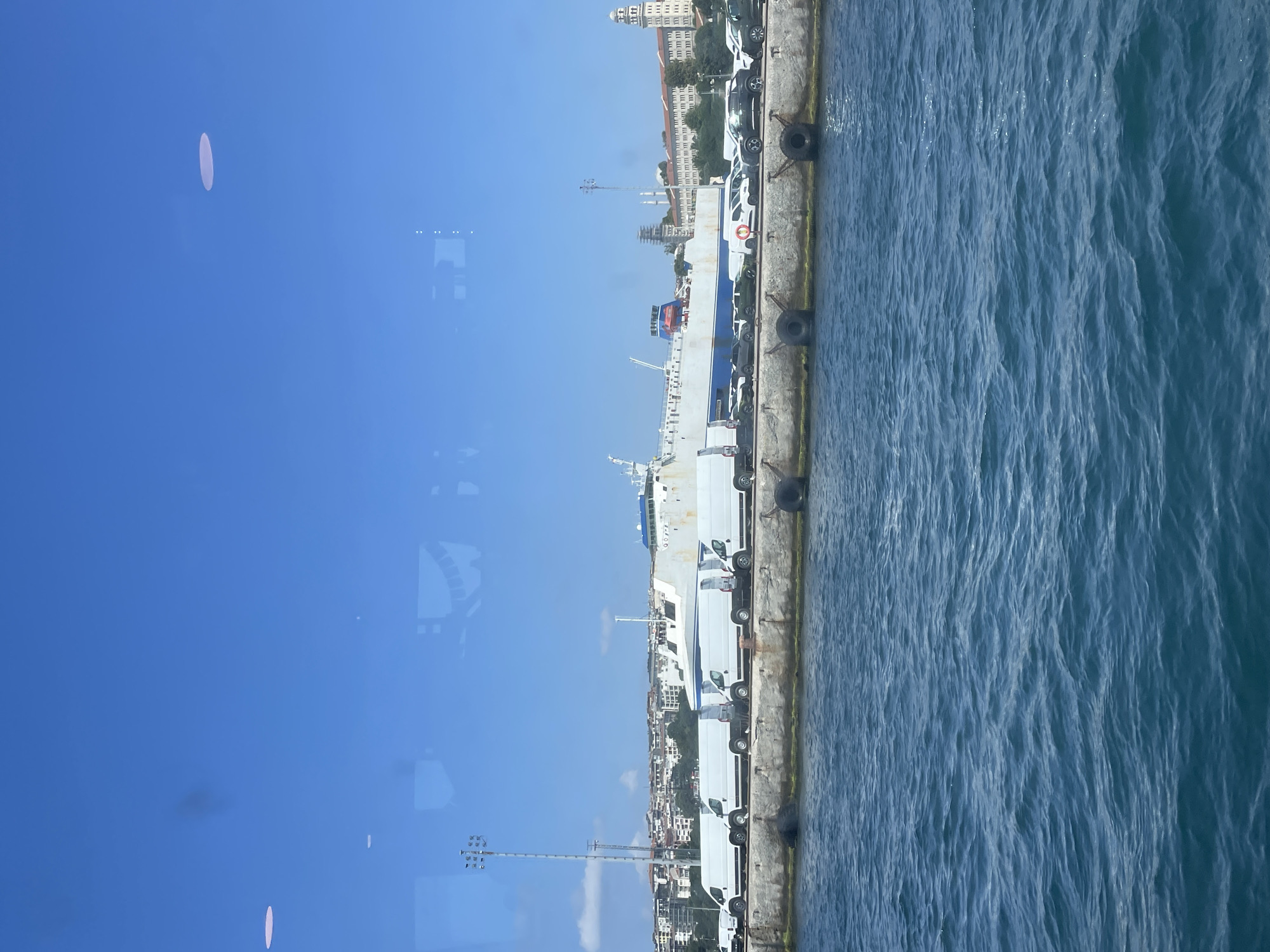 A long, white and blue ferryboat is docked at a port in Üsküdar, Turkey, under a clear blue sky. The ferry is filled with various white vans and cars, suggesting it is being used for transportation of vehicles. In the foreground, the blue water of the sea is rippling gently. The background shows a cityscape with buildings and trees. A tall light post stands on the left side of the image, and a circular white structure is visible in the sky on the left, possibly a reflection or a distant object. The overall scene depicts a bustling port activity on a sunny day.
