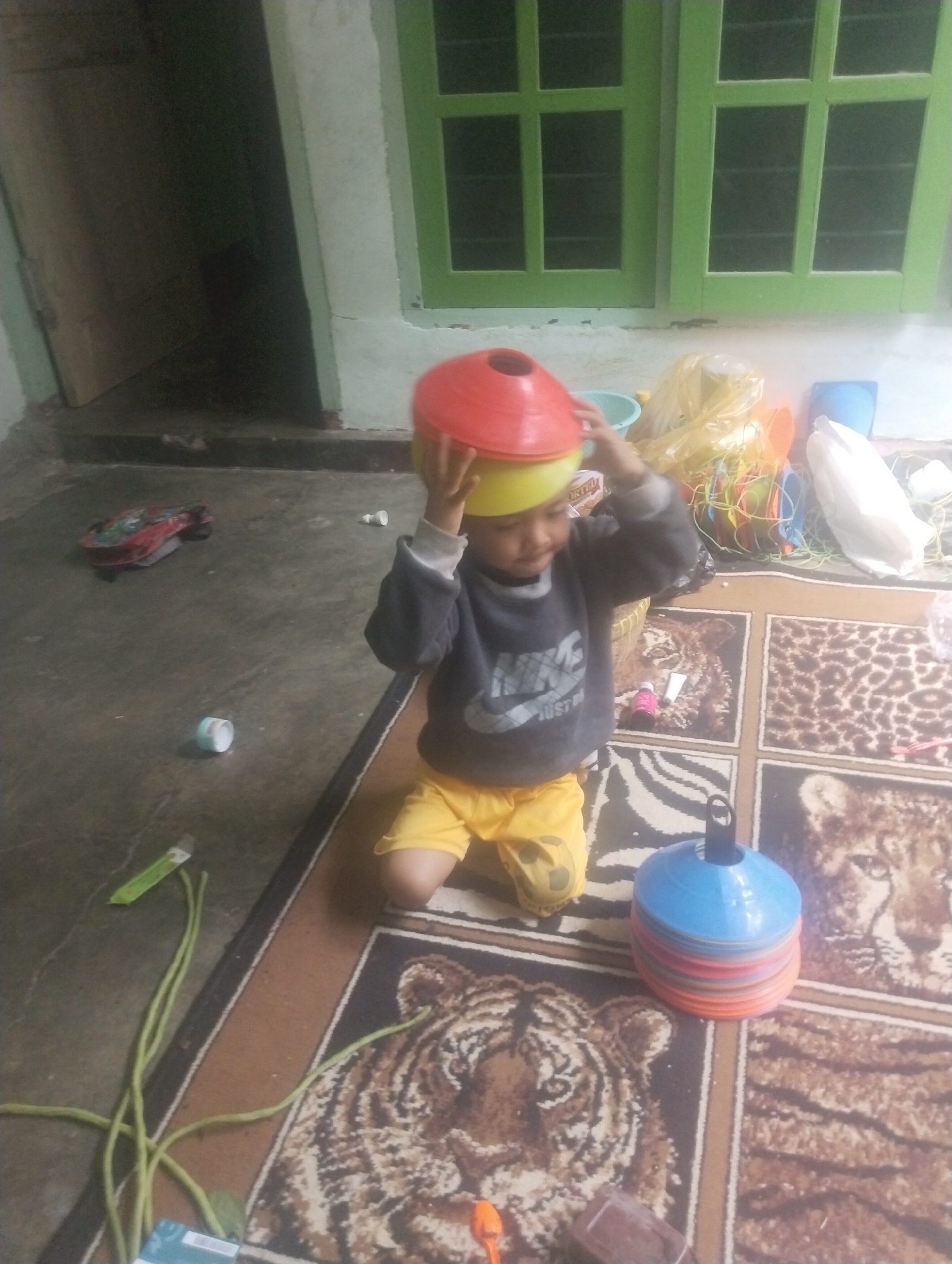 A young child, wearing a dark gray sweatshirt with a white "Nike" logo and bright yellow shorts, is playing indoors, balancing a yellow bowl on their head and a red cone on top of that. The child is kneeling on a rug with a tiger pattern. Scattered around the child are various toys and household items, including a stack of colorful cones, a yellow plastic bag, a small red toy car, and a bunch of long green vegetables. The background reveals a concrete floor, a doorway, and a window with green frames. The scene appears to be in a residential setting within Wanasaba Daya, Indonesia.