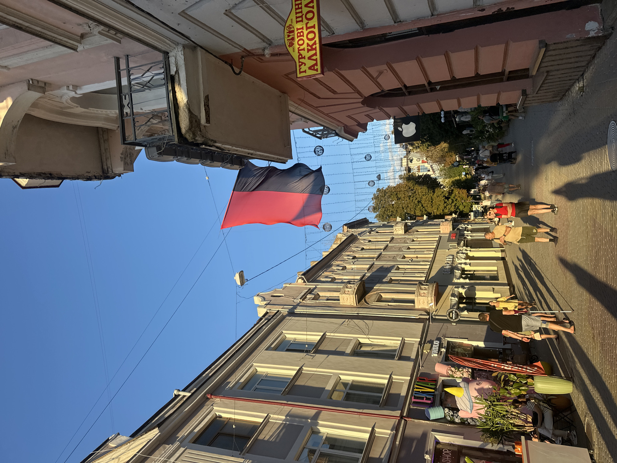 The image depicts a street scene in Ternopil, Ukraine, with architecture characteristic of Eastern Europe. The street is paved with cobblestones and lined with multi-story buildings. A red and black flag is prominently displayed, suspended from wires across the street. A yellow sign with Ukrainian text, "ГУРТОВІ ЦІНИ АЛКОГОЛЬ," which translates to "WHOLESALE PRICES ALCOHOL," is visible on the right side of the street.
Several people are walking along the street, some individually, others in small groups. One person is using a crutch. The sun is casting long shadows, indicating it is either early morning or late afternoon. Decorative string lights are hung across the street, suggesting a festive atmosphere or nighttime illumination. The buildings feature balconies and various architectural details. An Apple logo sign is visible hanging from one of the buildings further down the street. Lush greenery is present, with plants and trees interspersed along the sidewalk. The sky is a clear, bright blue.