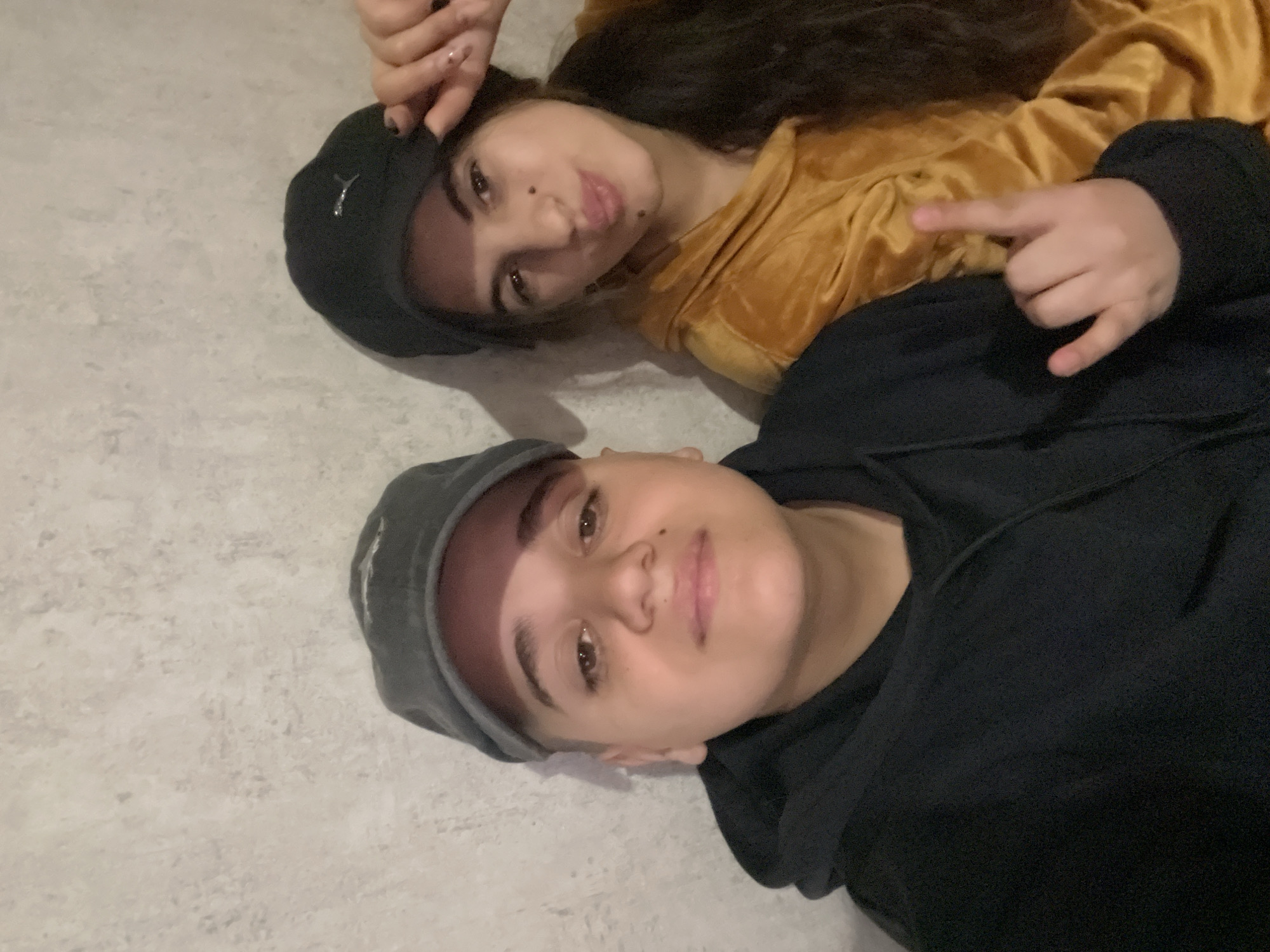 Two individuals are lying on a light-colored surface, likely a floor, indoors. The person in the foreground is wearing a dark grey cap and a black hooded sweatshirt. The second person, positioned behind and slightly above, is wearing a black baseball cap and a mustard-colored, velvet-like top. The individual in the back is making a "rock on" hand gesture. The photograph was likely taken in Lozarevo, Bulgaria.