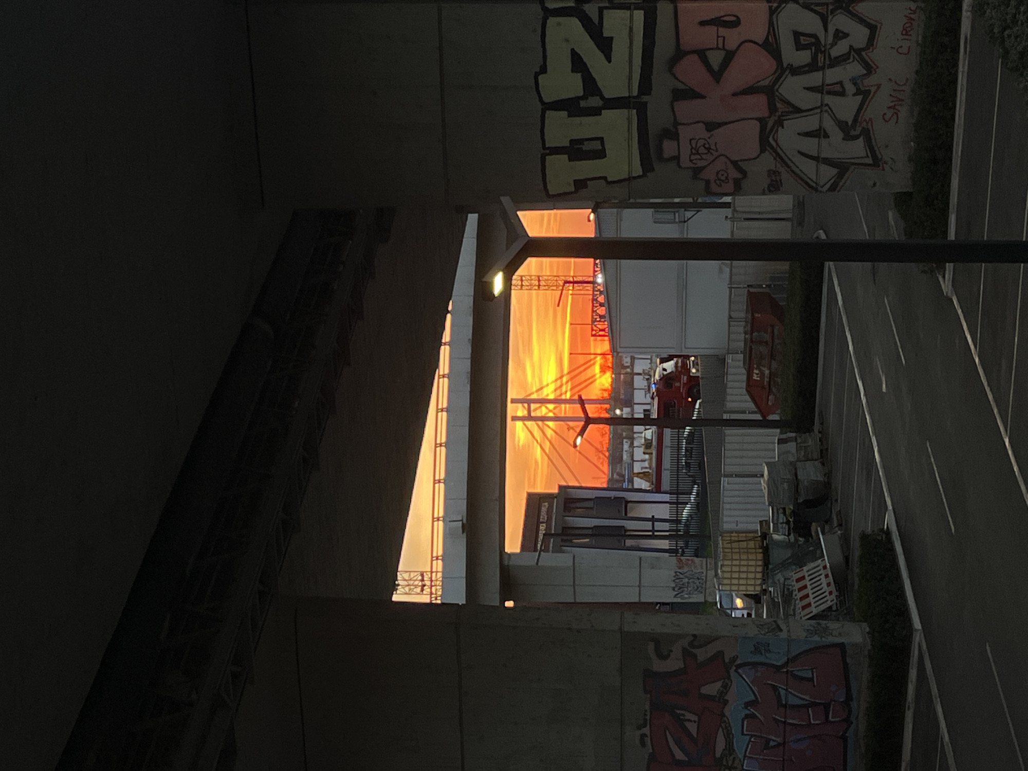 The image, taken from under a bridge in Belgrade, Serbia, shows a sunset or sunrise. The sky is orange, visible through the bridge's support structure. Graffiti adorns the bridge's walls. A parking lot with white lines is visible on the right side of the frame, and a few construction materials and bins are scattered near the edge. There is a firetruck or a vehicle resembling a firetruck, visible in the central view. A light post with an illuminated lamp is visible. The scene appears to be a mix of urban infrastructure, possibly an industrial area, and elements of city life.