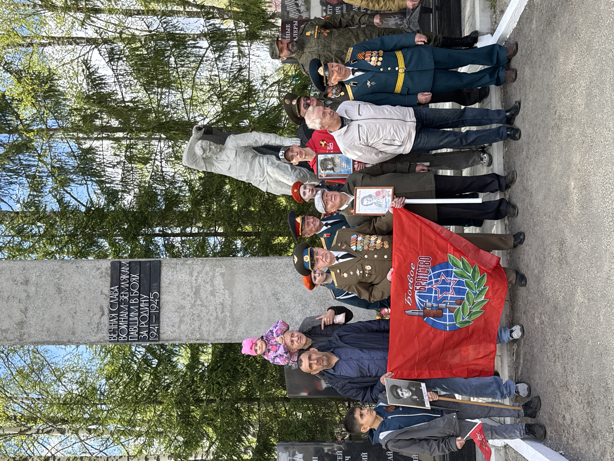 The image depicts a group of people gathered in what appears to be a memorial or commemorative event, likely related to World War II. The monument features inscriptions in Russian, "ВЕЧНАЯ СЛАВА ВОИНАМ-ЗЕМЛЯКАМ ПАВШИМ В БОЯХ ЗА РОДИНУ 1941-1945," which translates to "Eternal Glory to the soldiers-countrymen who fell in battle for the Motherland 1941-1945." There is also a statue and additional plaque that bears the city name as Yamyshevo, Russia.
Several individuals are holding portraits of soldiers, a red flag with a specific emblem, possibly representing a military organization or movement and miniature flags. Many of the people are wearing military uniforms or medals, indicating veterans or those associated with military service. The environment includes trees and what appears to be an outdoor memorial site or park. It is daytime and looks to be spring as the trees have new leaves.