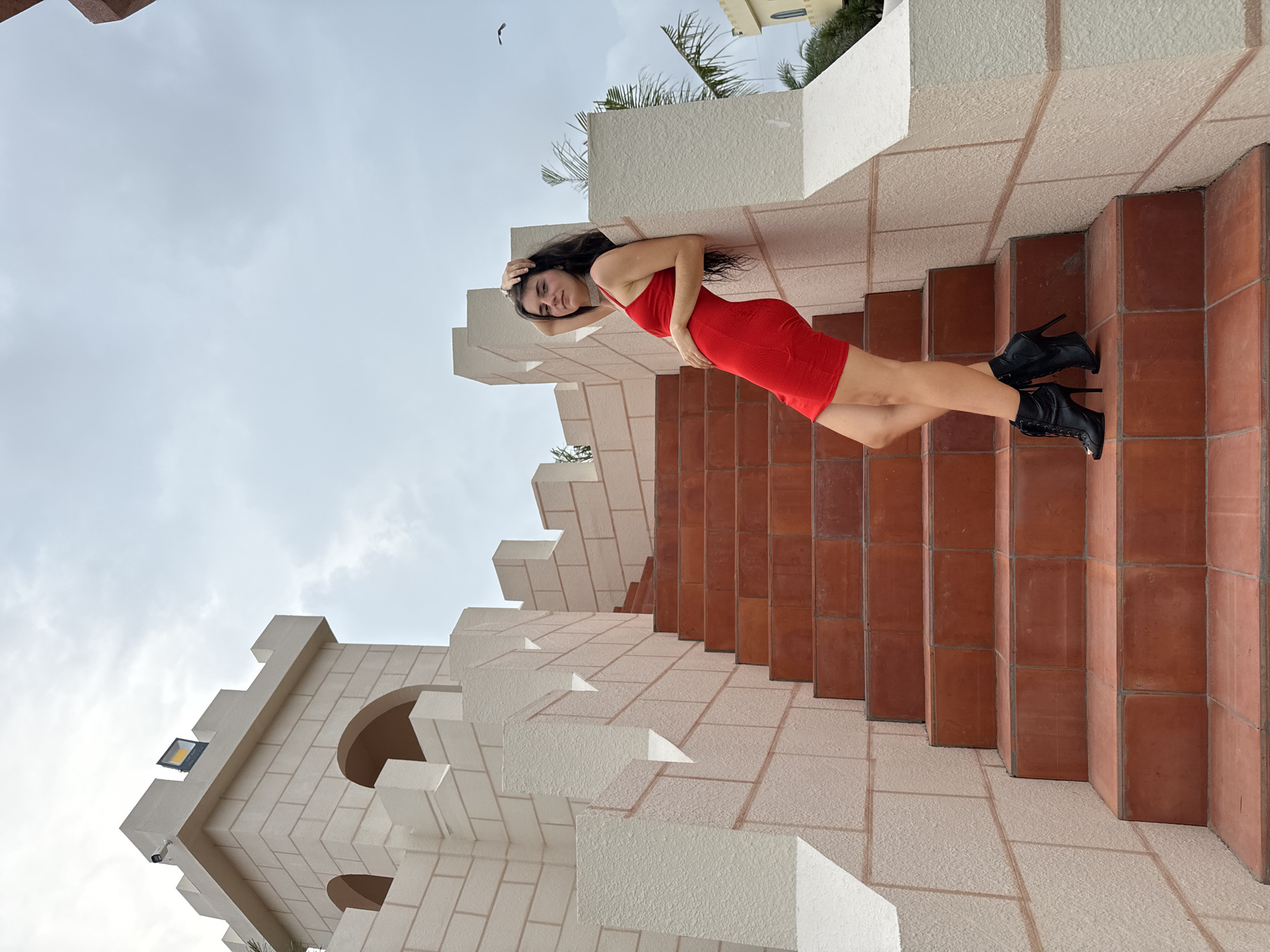 This media file captures a fashion-oriented outdoor portrait of a woman posing on a distinctive building, likely in Mui Ne, Vietnam. The image is oriented horizontally, with the woman and the architectural elements appearing to defy gravity, suggesting an intentional artistic rotation or a unique perspective.
The main subject is a woman with long, dark hair, wearing a form-fitting, sleeveless red mini-dress, black high-heeled lace-up ankle boots, and a sparkling choker necklace. She is leaning against a textured, cream-colored wall, looking towards the camera with a subtle expression. Her left hand rests on her hip, while her right hand is raised towards her head.
The setting is characterized by elaborate, castle-like architecture. The building features light cream or off-white walls with prominent crenellated sections and block patterns, along with arched openings. A set of broad stairs or terraced platforms, made of reddish-brown square tiles, ascends from the lower part of the frame towards the right. The sky overhead is overcast and light grey. In the background, hints of other buildings and lush palm trees are visible, along with a small bird flying high in the sky. A practical detail on one of the castle-like structures includes a mounted spotlight or floodlight and a small black security camera.
The overall scene suggests a stylish photoshoot taking place during the daytime under cloudy conditions, utilizing the unique architectural lines and textures of the location. There is no visible text in the image.