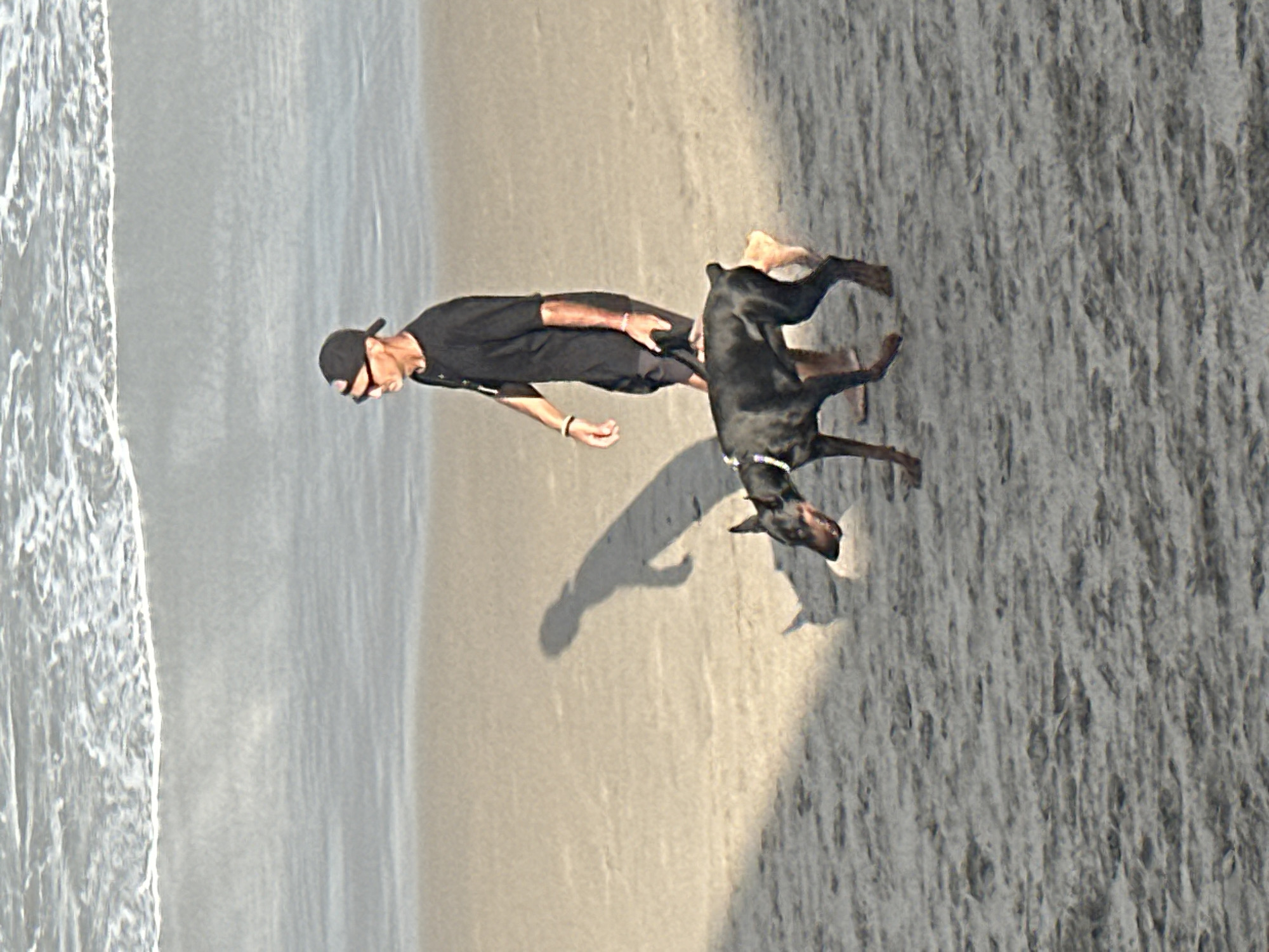 A man and a Doberman Pinscher are walking on a sandy beach. The man is wearing a black t-shirt, black shorts, a black baseball cap, and sunglasses. He has a leash in his right hand attached to the dog's collar. The dog is black and tan and appears to be walking ahead of the man. The background consists of the ocean with waves breaking on the shore. The scene is set during the daytime, as indicated by the shadows cast on the sand. The location appears to be a beach in North Kuta, Indonesia.